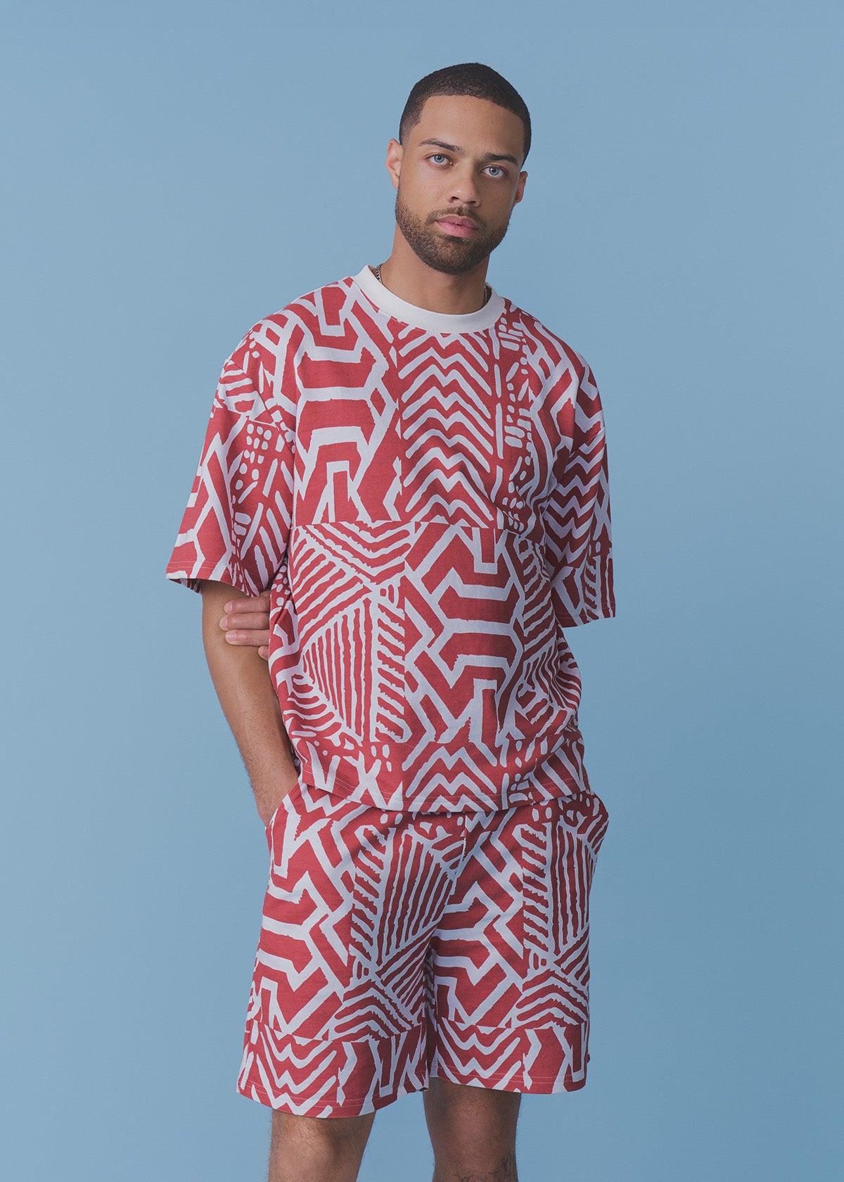 A man modeling a matching short-sleeve t-shirt and shorts set with a bold red and white geometric maze-like pattern, set against a solid light blue background.