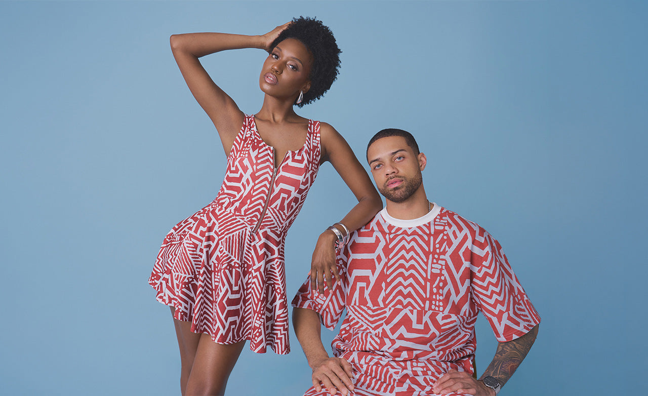 A man and a woman modeling matching red and white patterned clothing against a solid blue studio background. The woman wears a sleeveless red and white geometric print mini dress, and the man wears a matching red and white patterned short-sleeve top and matching bottoms.