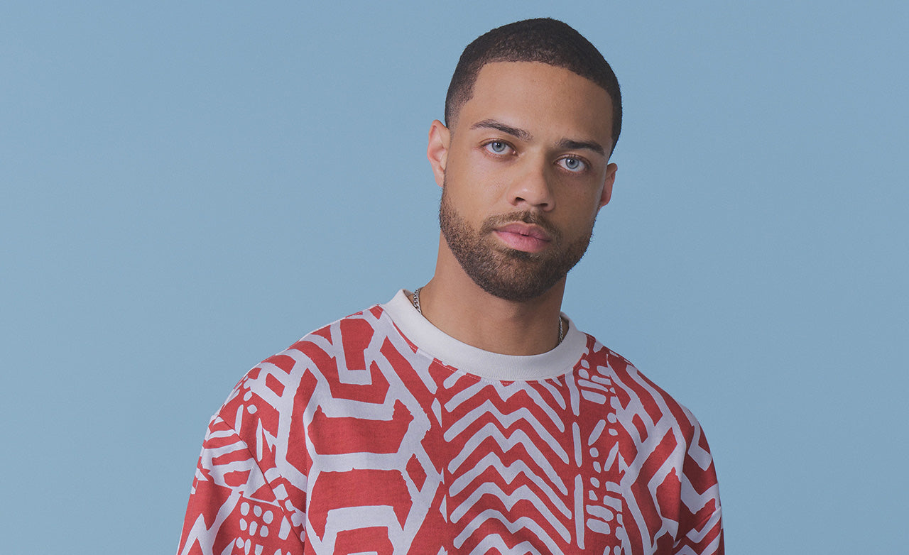 A man with short dark hair and a beard wearing a red and white patterned crewneck sweatshirt, photographed against a solid light blue background.