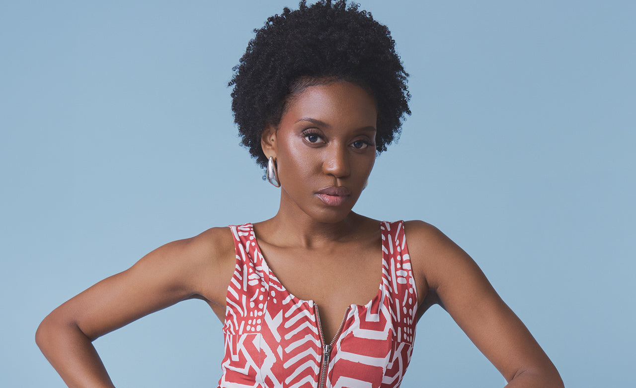 A woman with short curly black hair wearing a red and white geometric patterned sleeveless top, posing against a solid light blue background.
