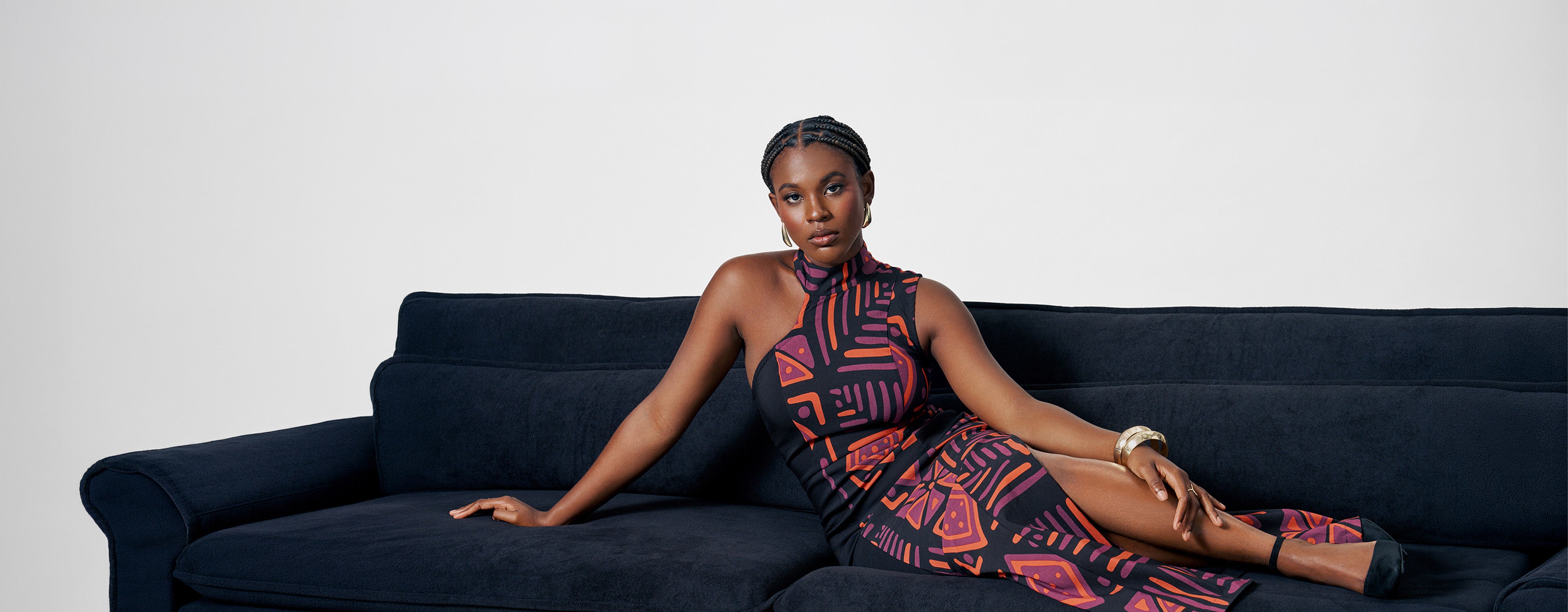 Woman reclining on navy sofa wearing sleeveless black midi dress with purple and orange African inspired geometric print