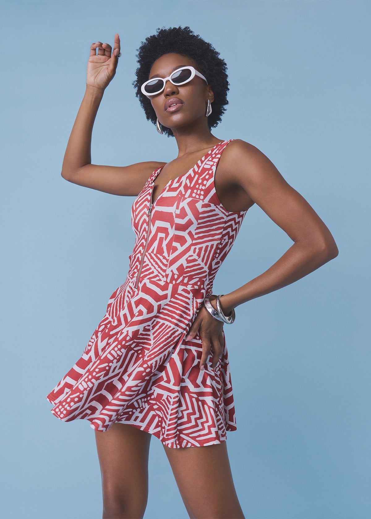 A woman modeling a sleeveless V-neck mini dress with a red and white geometric pattern, styled with white sunglasses, against a solid light blue background.