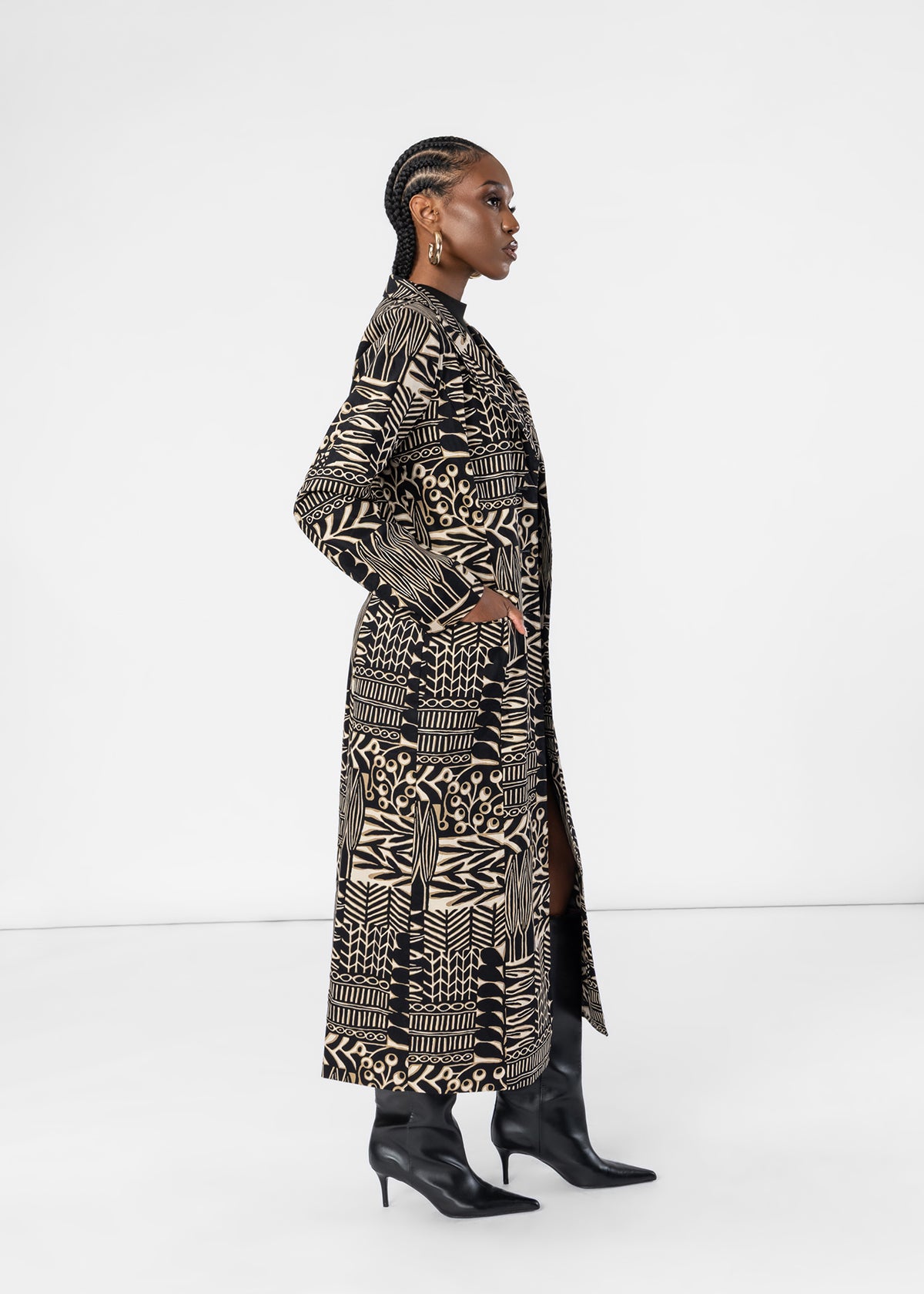 A woman modeling a long black and beige geometric patterned coat with black pointed-toe boots, shown in profile against a plain white studio background.