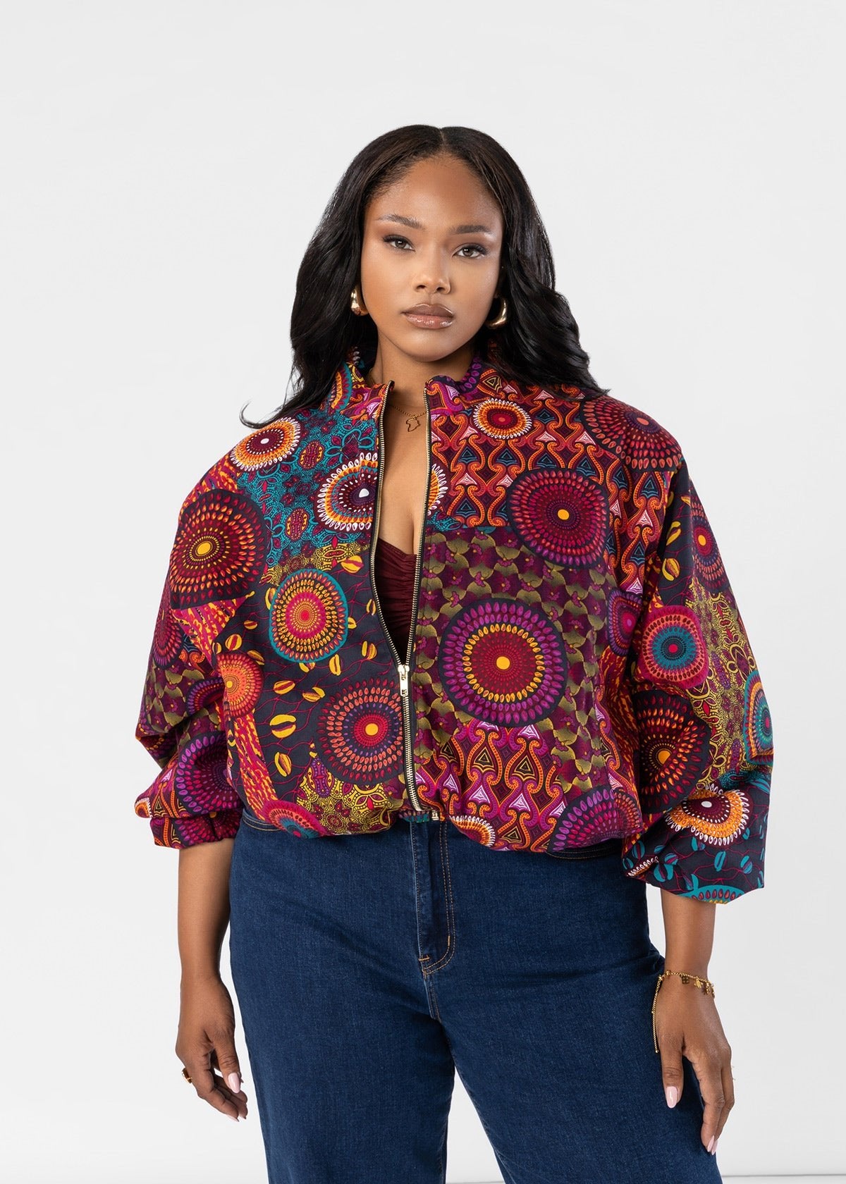 A woman modeling a vibrant, multicolored patterned bomber jacket with balloon sleeves, paired with dark blue jeans, against a plain white studio background.