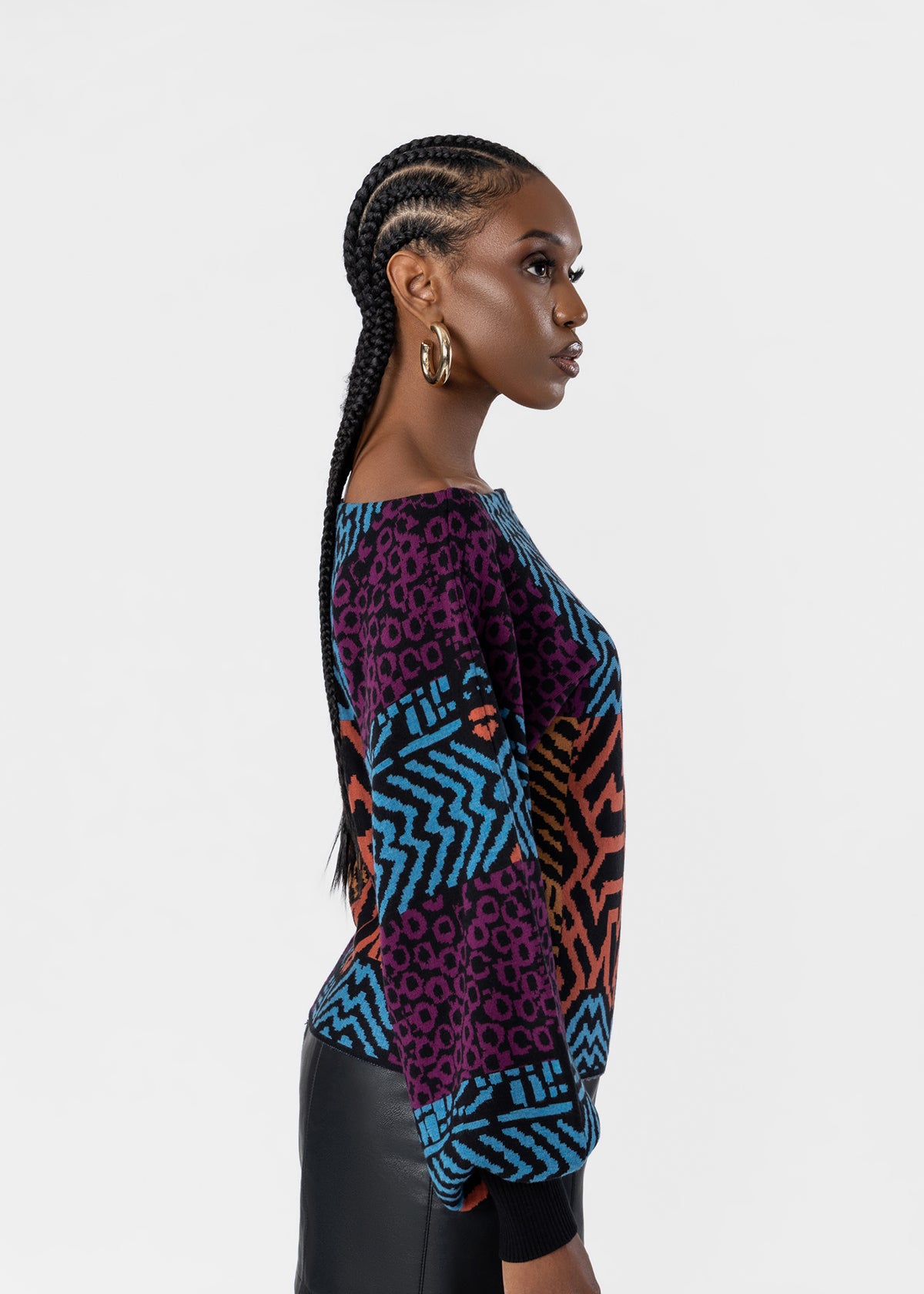 A woman in profile wearing a colorful patterned knit top with purple, blue, and orange geometric and leopard-print designs, paired with black bottoms, against a light gray studio background.