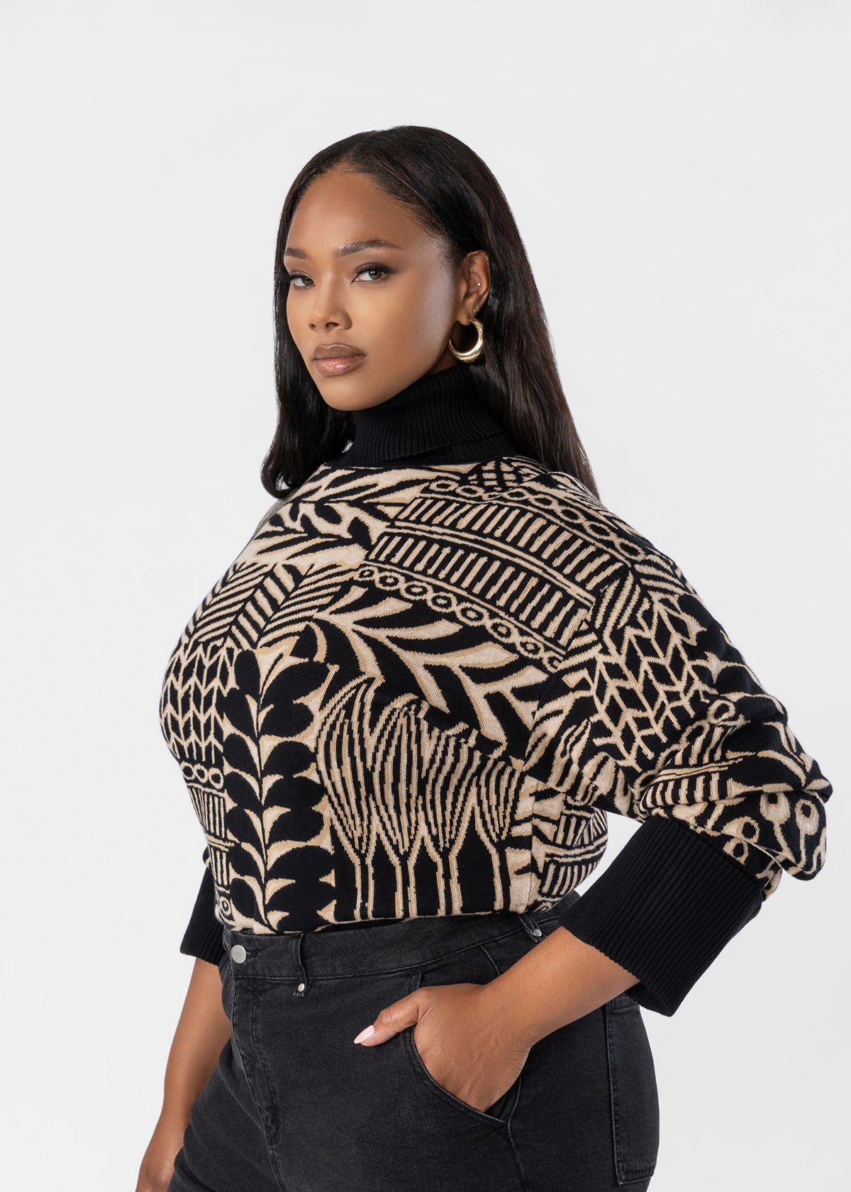 A woman modeling a cropped knit sweater with a bold black and beige botanical pattern, paired with black jeans against a light gray studio background.