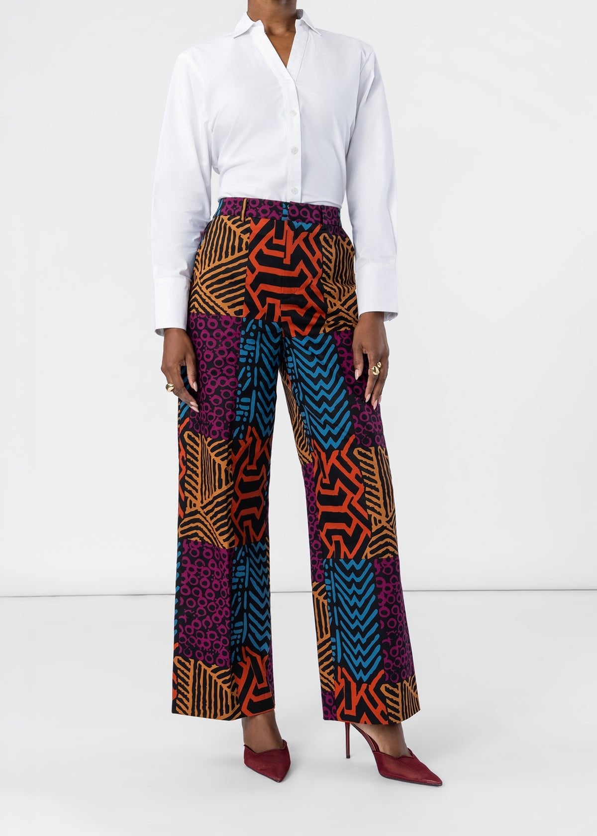 A model wearing a white button-down shirt and high-waisted, wide-leg trousers featuring a vibrant multicolor geometric and animal-print patchwork pattern in orange, blue, purple, and black. The model is also wearing burgundy pointed-toe heels against a clean white studio background.
