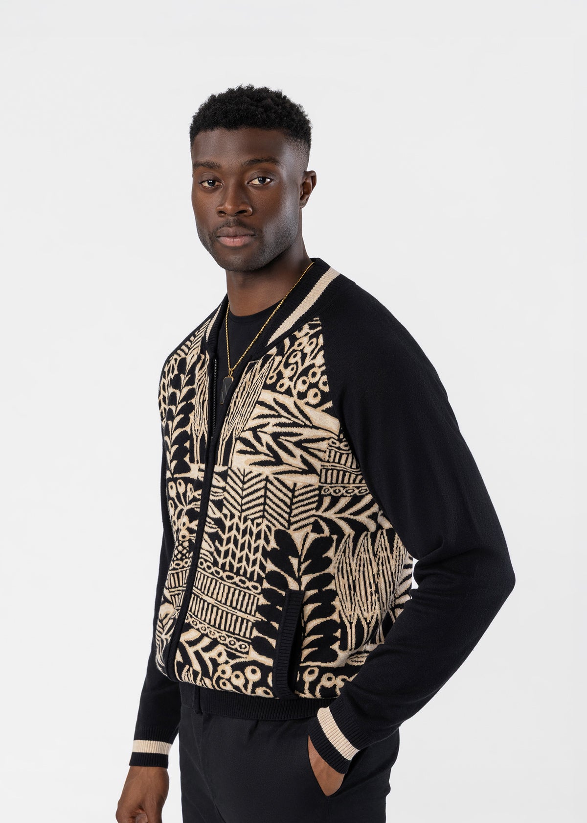 A man wearing a black and beige patterned zip-up bomber jacket with solid black sleeves, standing against a light gray studio background.