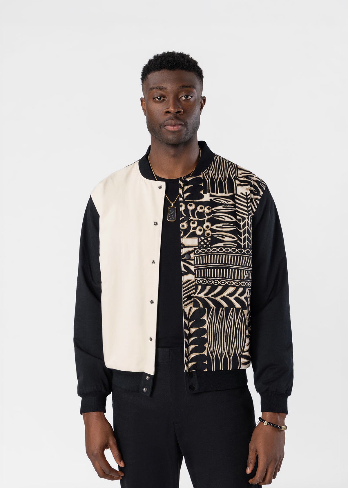 A man wearing a black and cream color-blocked bomber jacket with a detailed black and cream geometric pattern on the right side, paired with black pants against a light gray background.