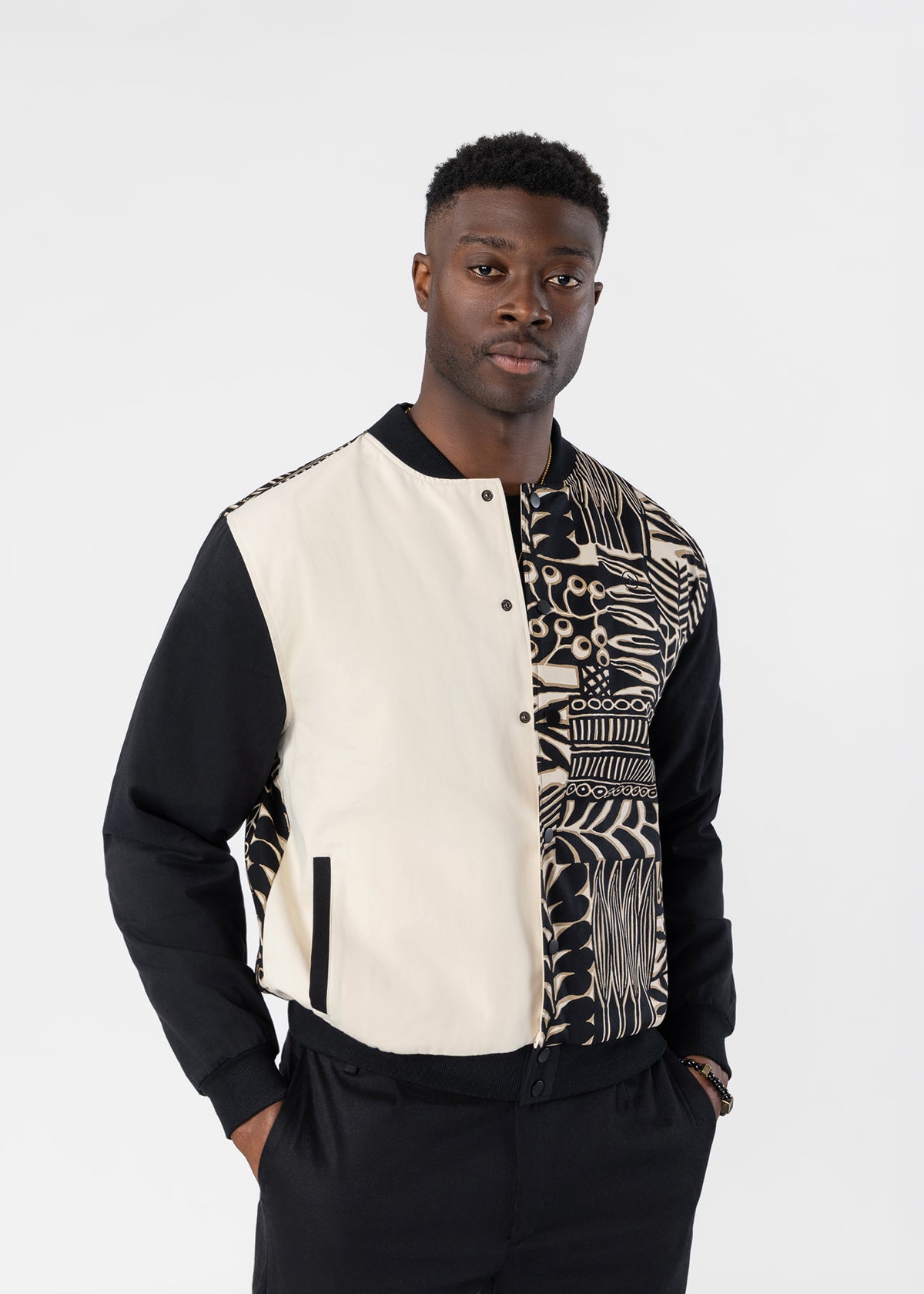 A man wearing a modern color-blocked bomber jacket with black sleeves, a cream center panel, and a black-and-white geometric patterned panel on the right side, posed against a light gray studio background.