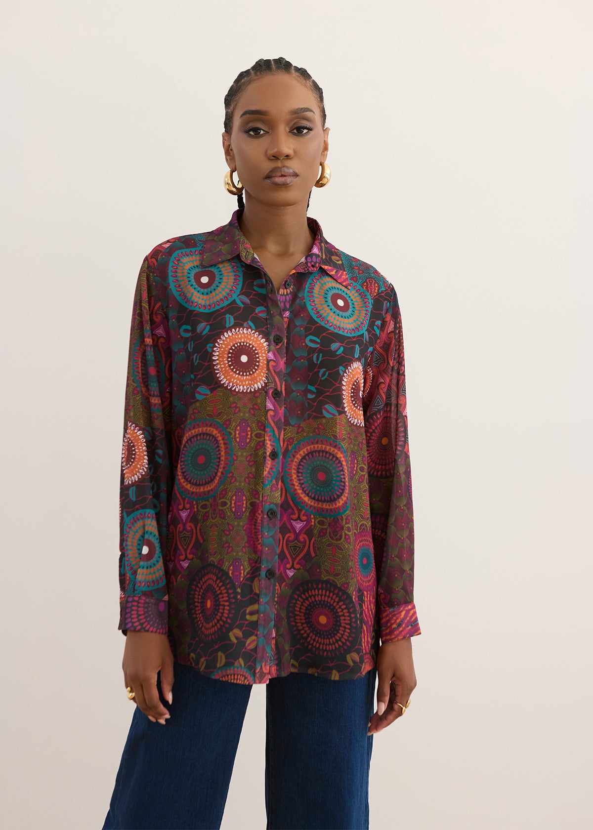A woman modeling a long-sleeve button-up shirt with a vibrant, multicolored circular mandala pattern in red, orange, blue, and green on a dark base, paired with dark blue jeans against a plain light beige background.