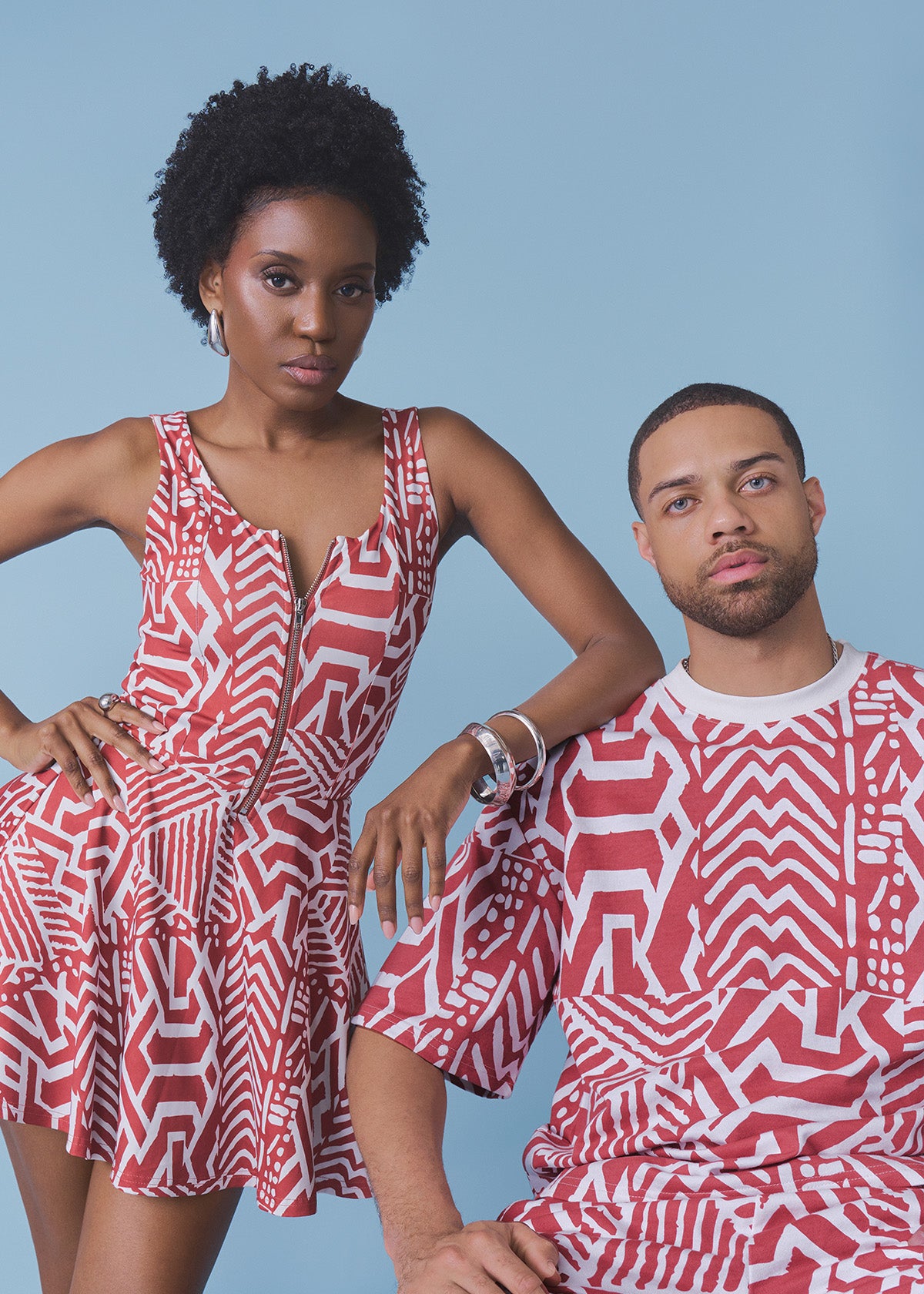 A man and a woman wearing matching red and white geometric patterned outfits, posed against a solid light blue background.
