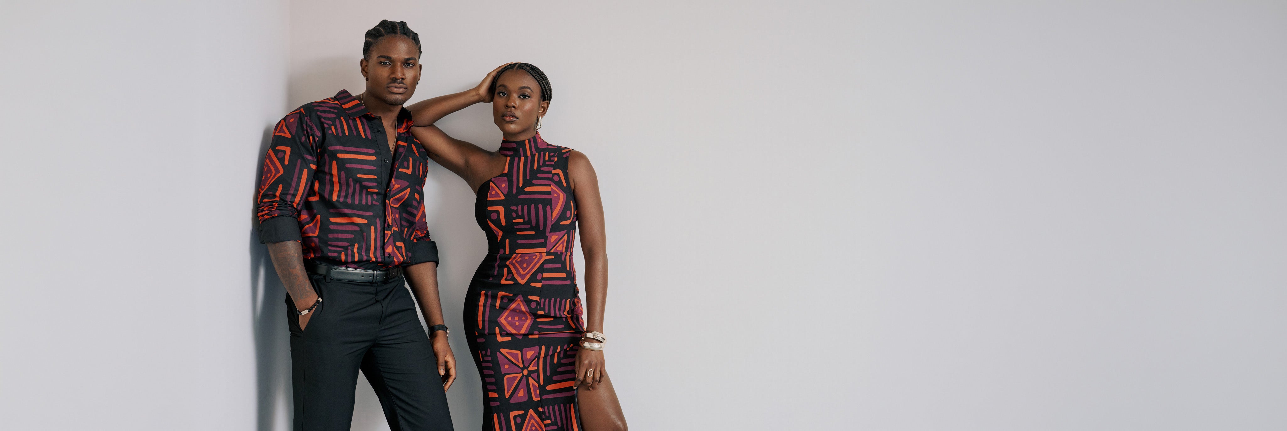 His and hers models wearing matching black outfits with red-orange African-inspired print against neutral studio wall