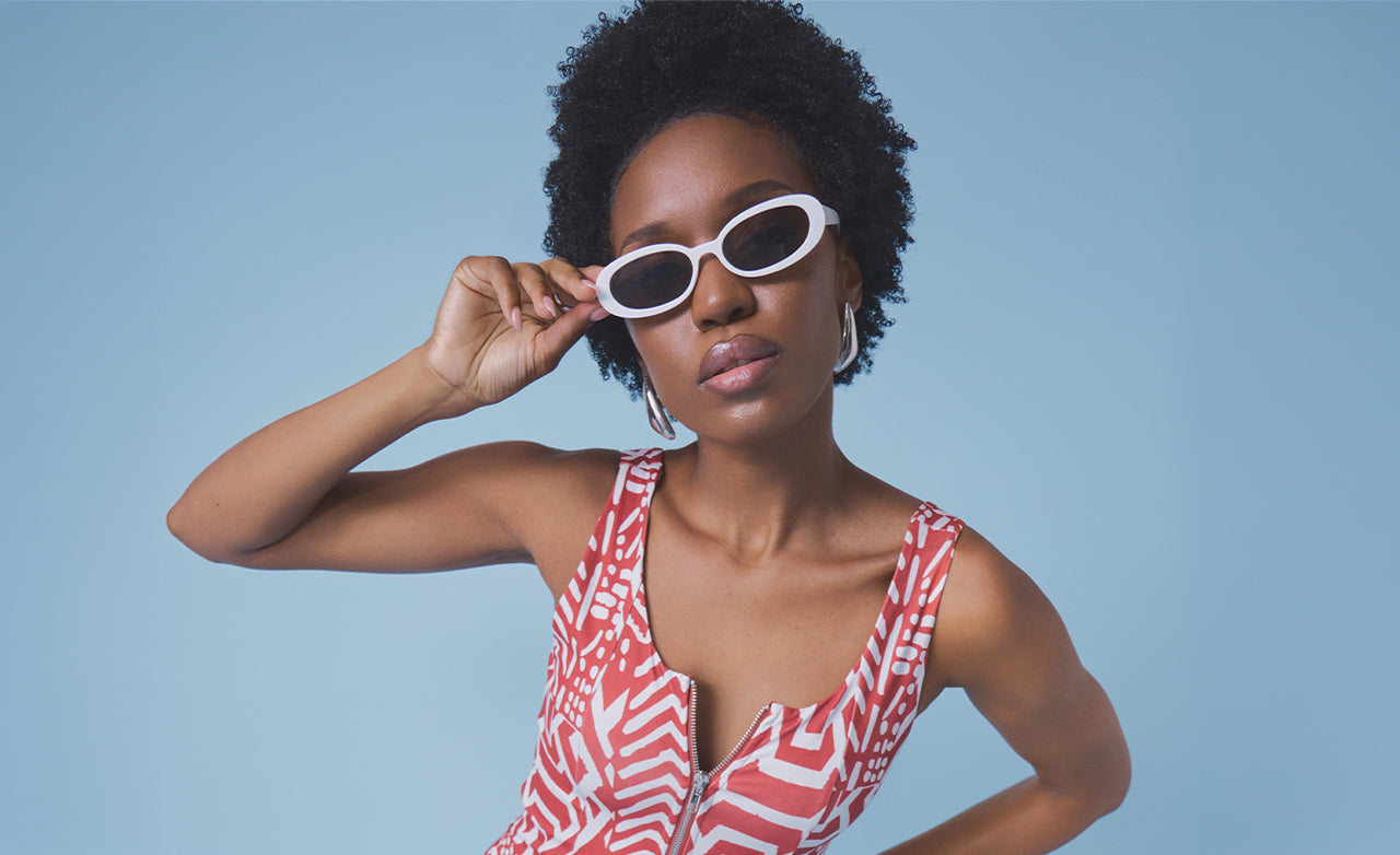A woman with short curly black hair wearing white oval sunglasses and a red and white patterned top, posing against a solid light blue background.