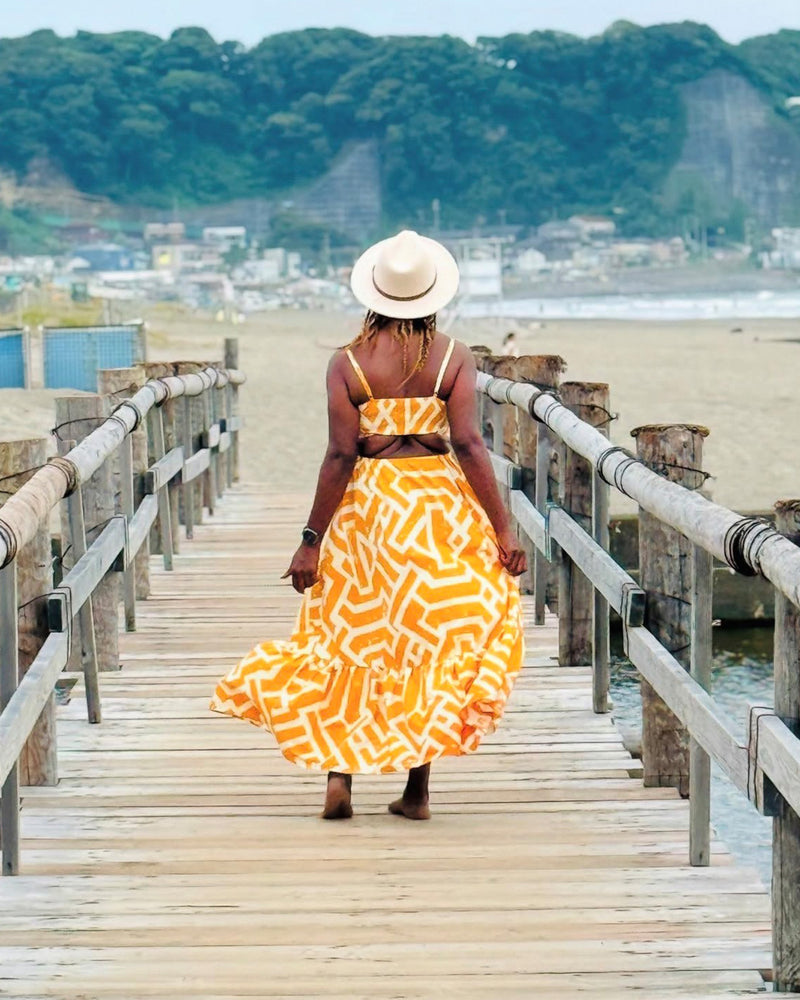 Woman in a gold and cream African print maxi dress walks along a wooden pier toward a beach with hills