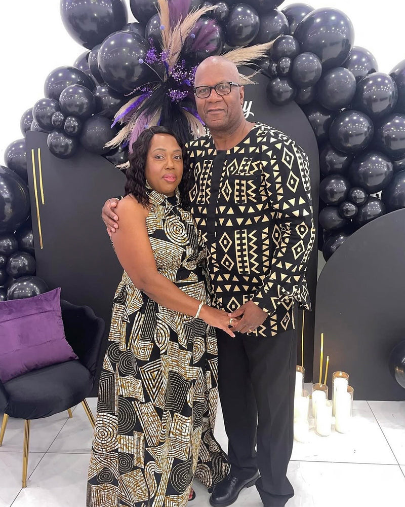 Woman models a black and brown geometric halter maxi dress beside a man in patterned clothing in front of a black balloon display