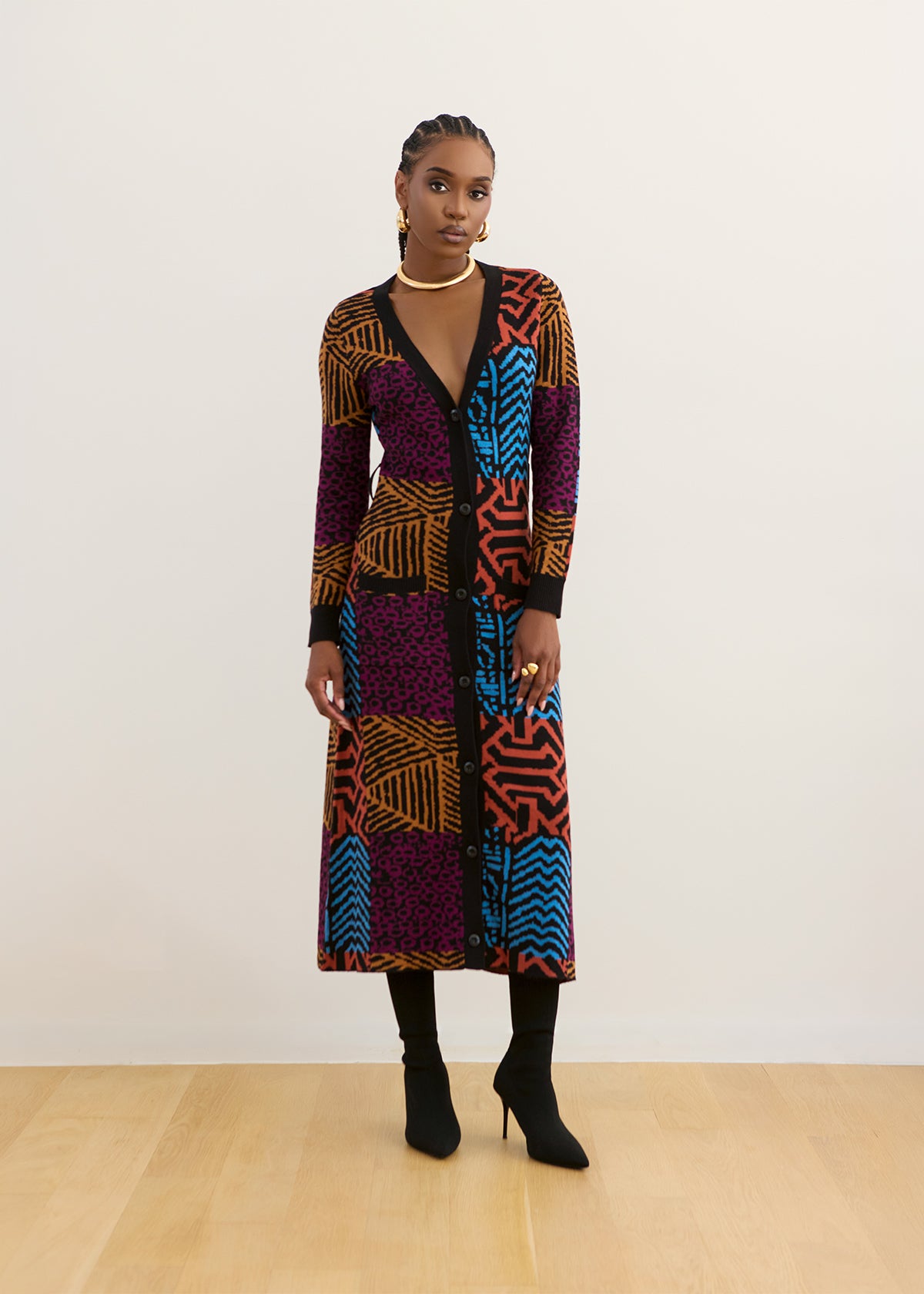 A model stands in a studio wearing a long-sleeve midi wrap dress with a vibrant multicolor geometric and abstract patchwork pattern in purple, orange, blue, and black. She is also wearing black knee-high socks and black high-heeled boots against a plain off-white background with light wood flooring.