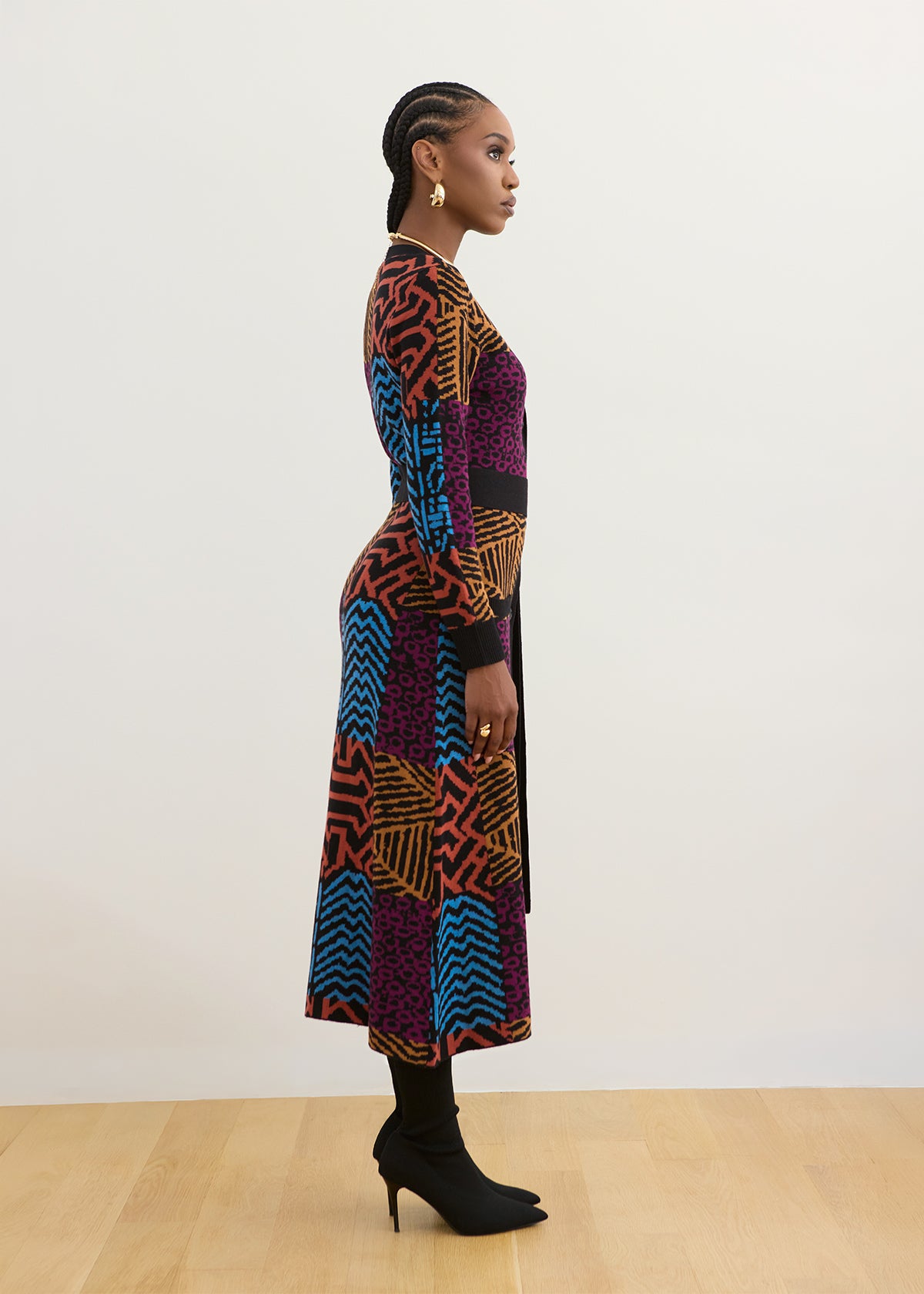 A woman stands in profile wearing a long-sleeve midi dress with a bold multicolor geometric and abstract print in purple, blue, orange, black, and red, paired with black tights and black ankle boots against a plain off-white wall and light wood floor.