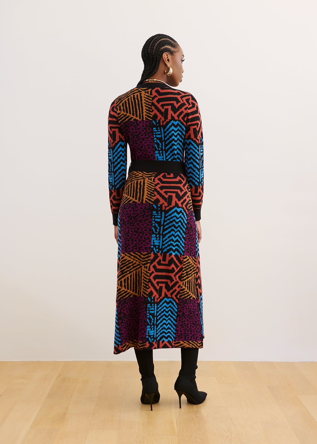 A model is shown from the back wearing a long-sleeved midi dress with a vibrant multicolor geometric patchwork pattern in purple, blue, orange, and red, paired with black boots and a black belt, set against a minimalist white wall and light wood floor.