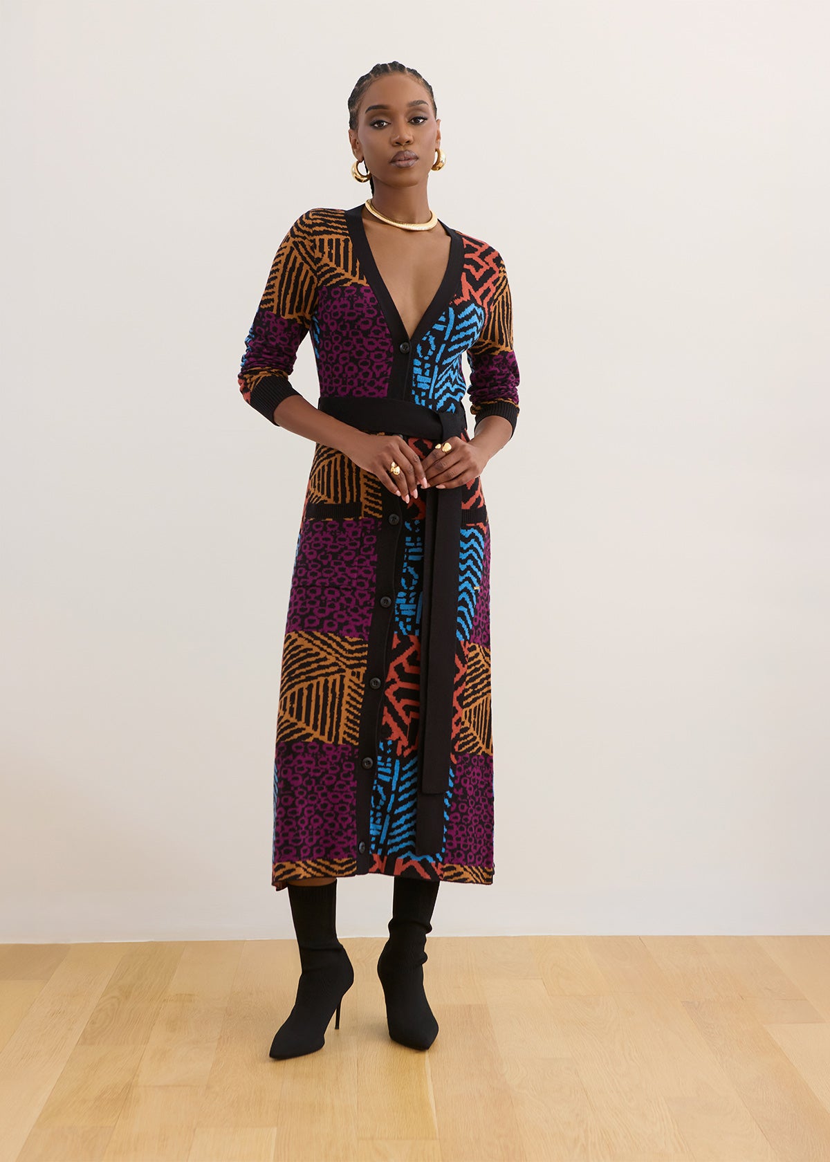A model stands in a studio wearing a long-sleeve wrap dress with a vibrant multicolor geometric and leopard print pattern in purple, blue, orange, and black. She is also wearing black ankle boots, black tights, and gold jewelry against a plain white wall and light wood floor.