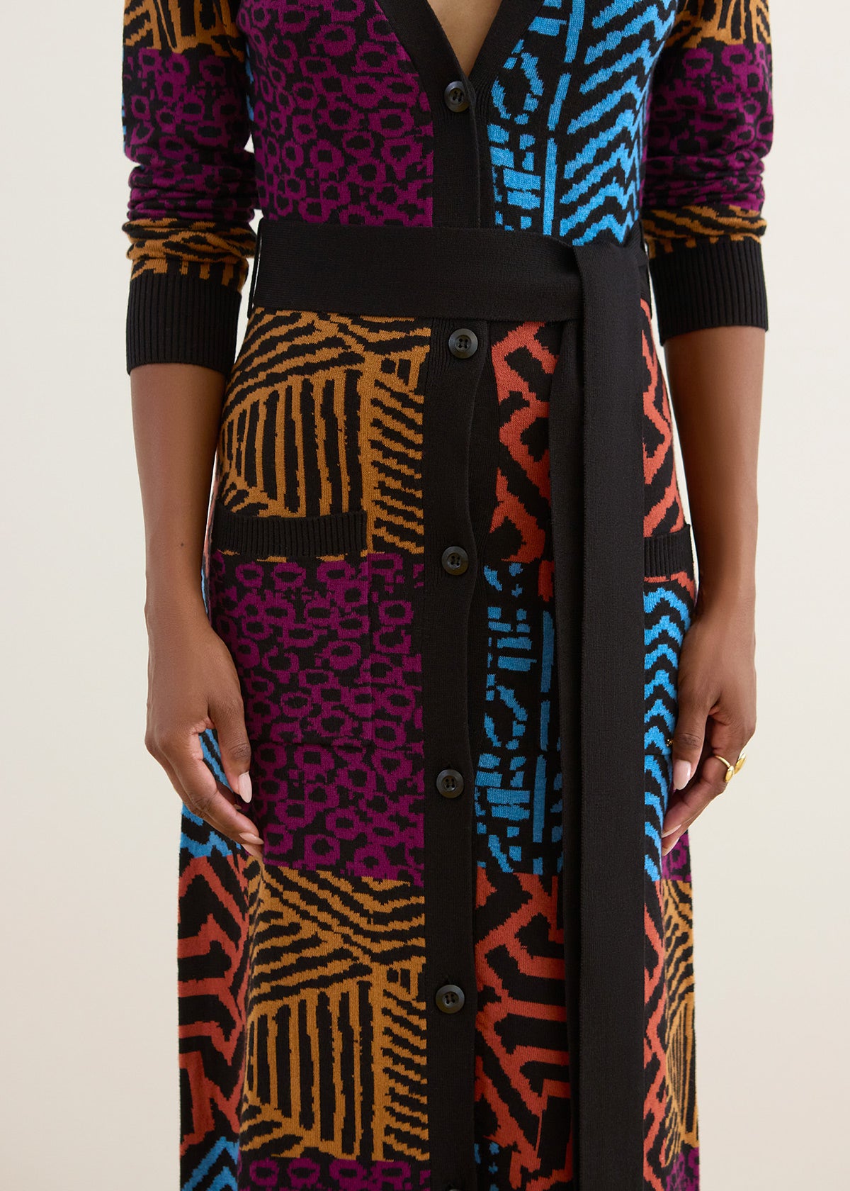 A close-up of a model wearing a long-sleeve button-front cardigan dress with a bold multicolor geometric and animal print pattern, featuring black ribbed trim and a matching black waist belt.