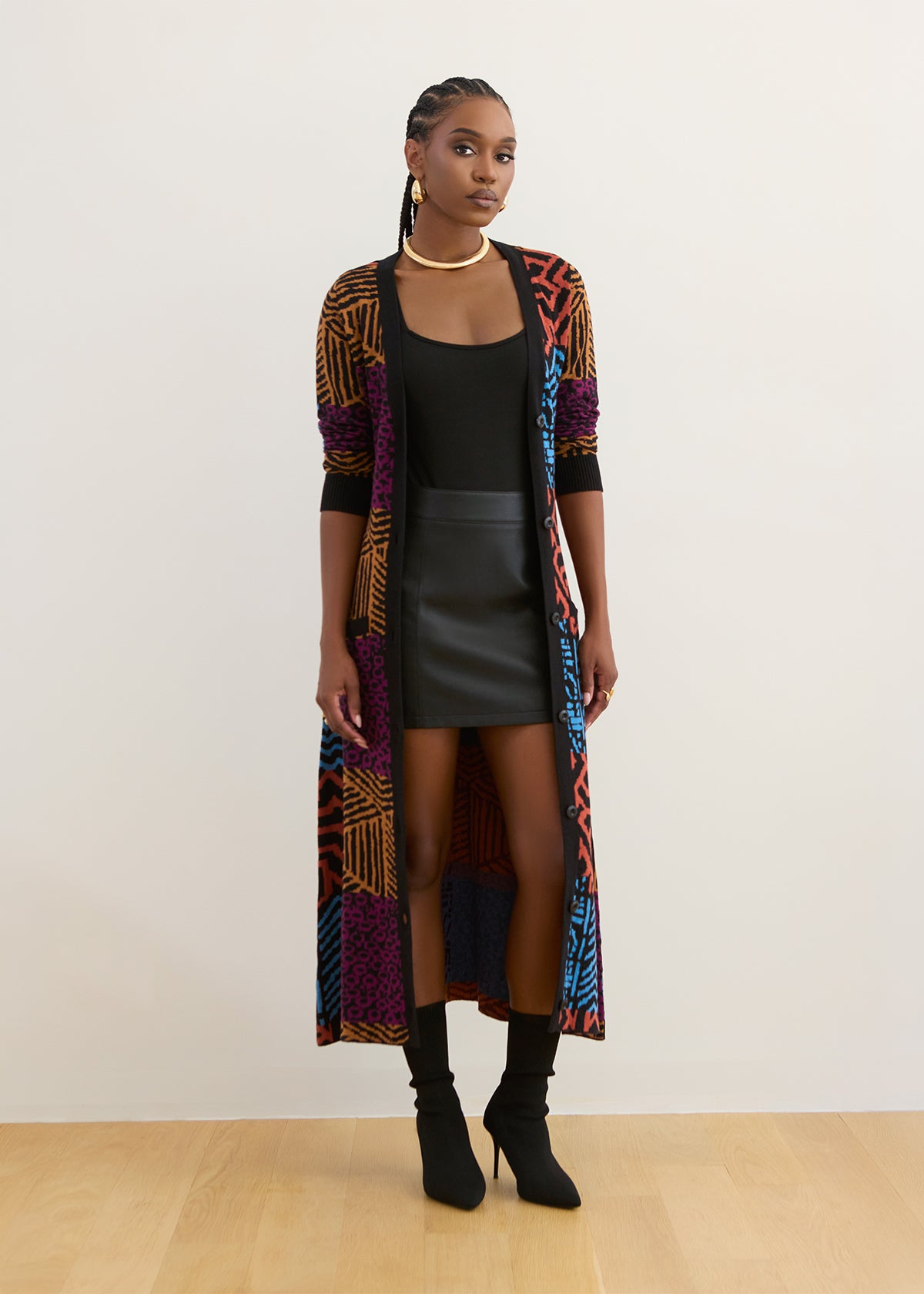 A model stands against a plain off-white wall wearing a long open-front cardigan with a vibrant multicolor geometric pattern, styled over a black top and a black faux-leather mini skirt.