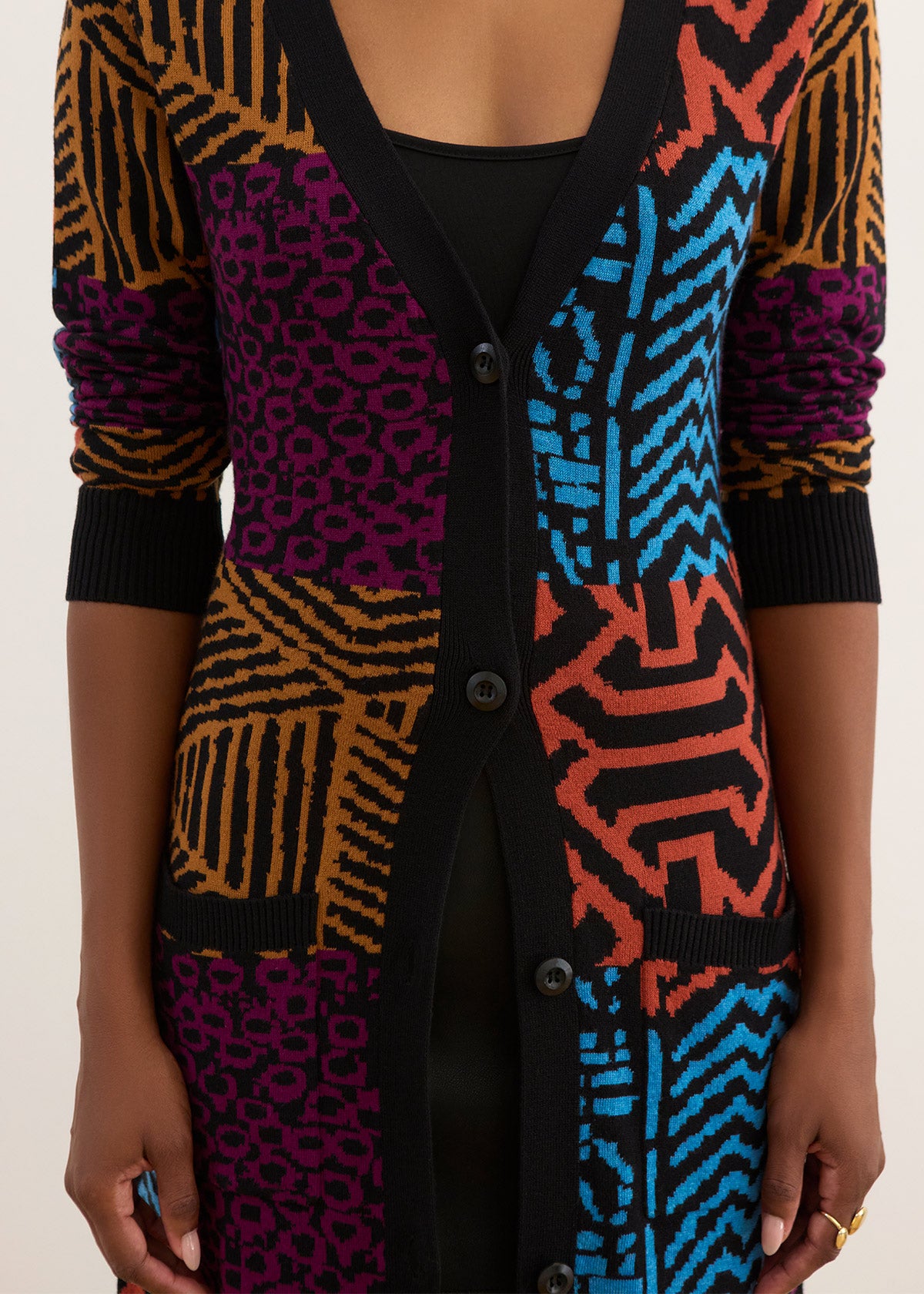 A close-up of a person wearing a long open-front cardigan with a bold multicolor patchwork knit pattern. The cardigan features sections of black, bright blue, orange, and purple with geometric and animal-print motifs, worn over a black top against a plain light background.