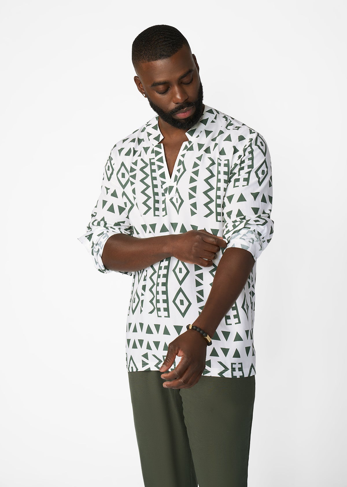 A man wearing a white long-sleeve button-up shirt with a dark green geometric tribal pattern, paired with olive green trousers, photographed against a plain white studio background.