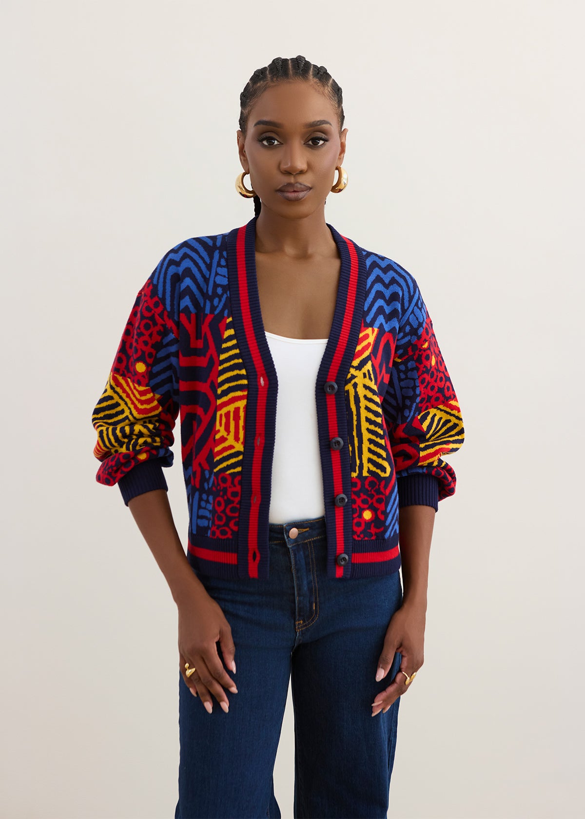A woman modeling a vibrant, multicolored African wax print cardigan with red, blue, yellow, and black patterns, styled over a white tank top and dark blue jeans against a plain light background.