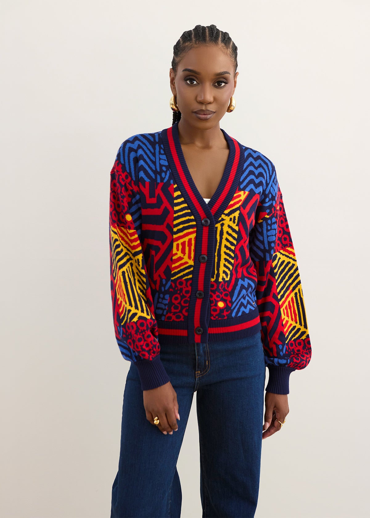 A model wearing a vibrant, multicolored geometric patterned cardigan with blue, red, yellow, and orange panels, paired with dark blue jeans, against a plain light gray background.