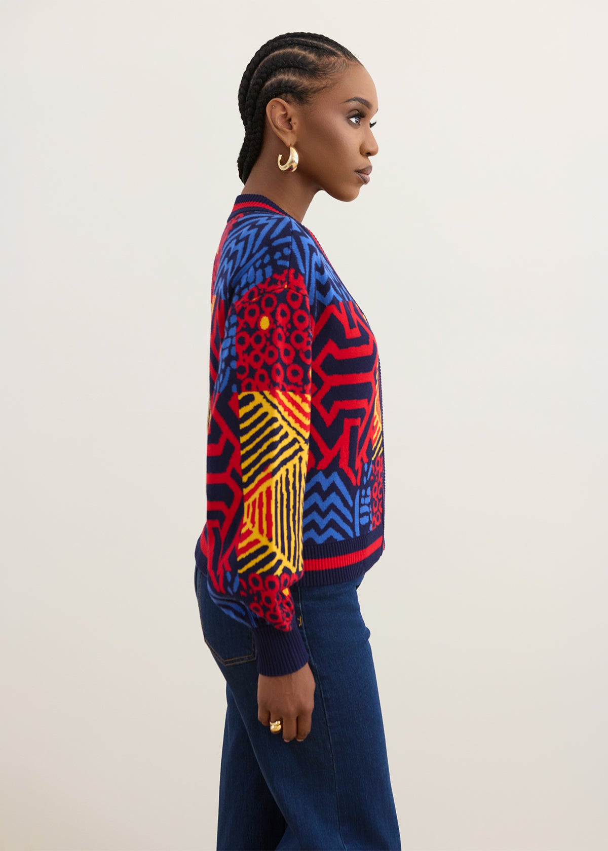 A woman in profile wearing a vibrant, multicolored geometric patterned sweater and dark blue jeans, standing against a plain light beige studio background.