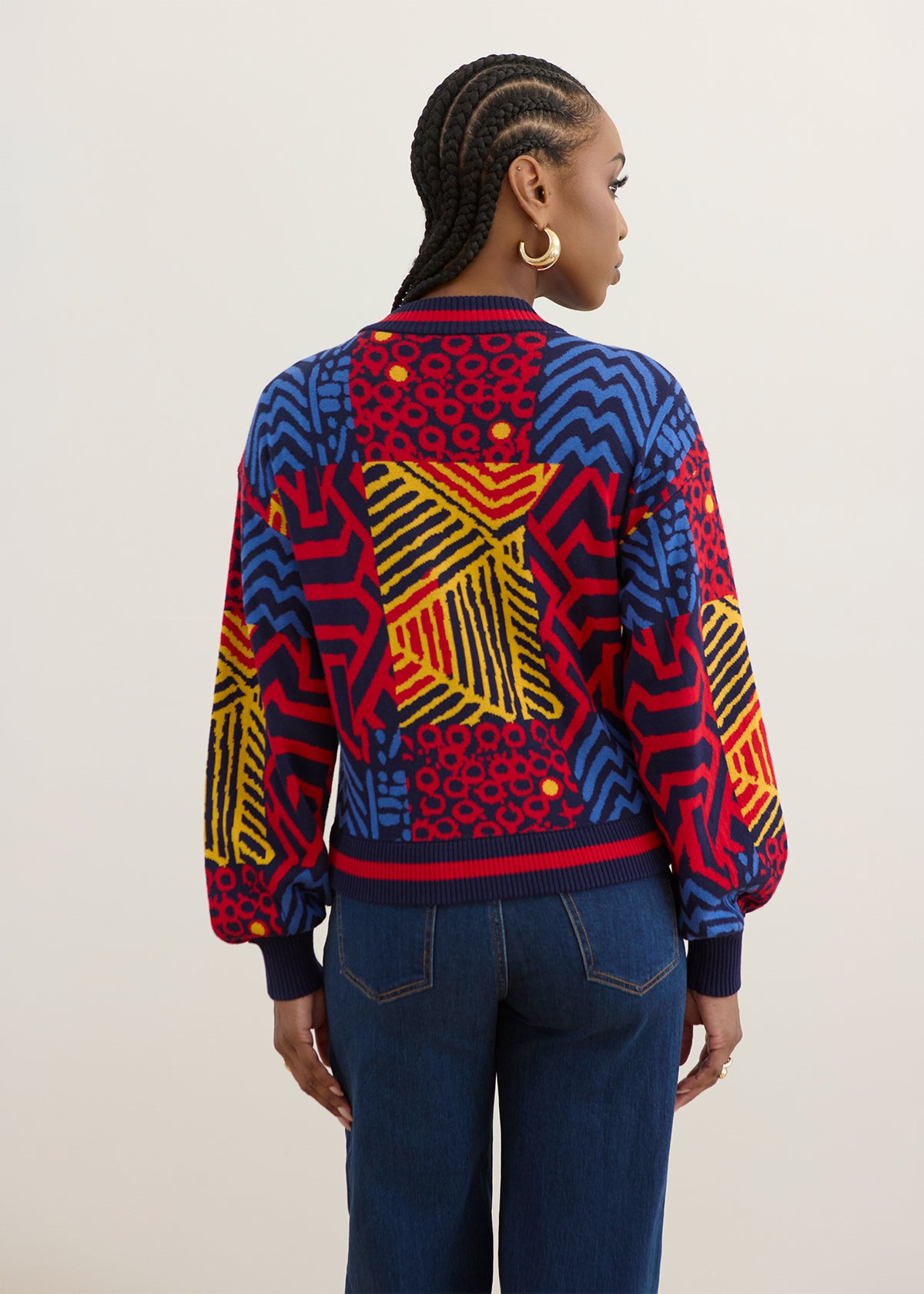 A model shown from the back wearing a vibrant, multicolor patterned knit bomber jacket with blue, red, yellow, and black geometric designs, paired with dark blue jeans against a plain light gray background.