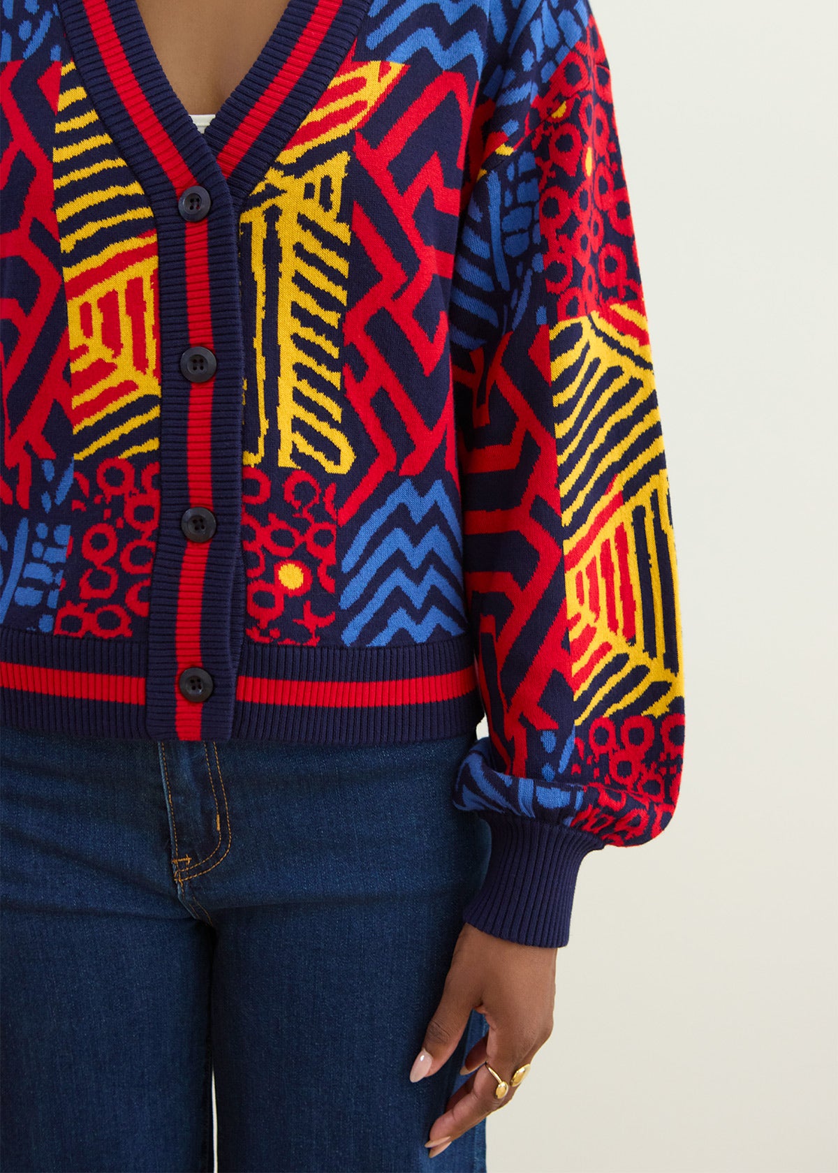 A close-up of a person wearing a colorful geometric patterned cardigan sweater with blue jeans. The cardigan features a navy blue ribbed V-neck, button placket, cuffs, and hem, with vibrant red, yellow, and blue patterned panels on the body and sleeves.