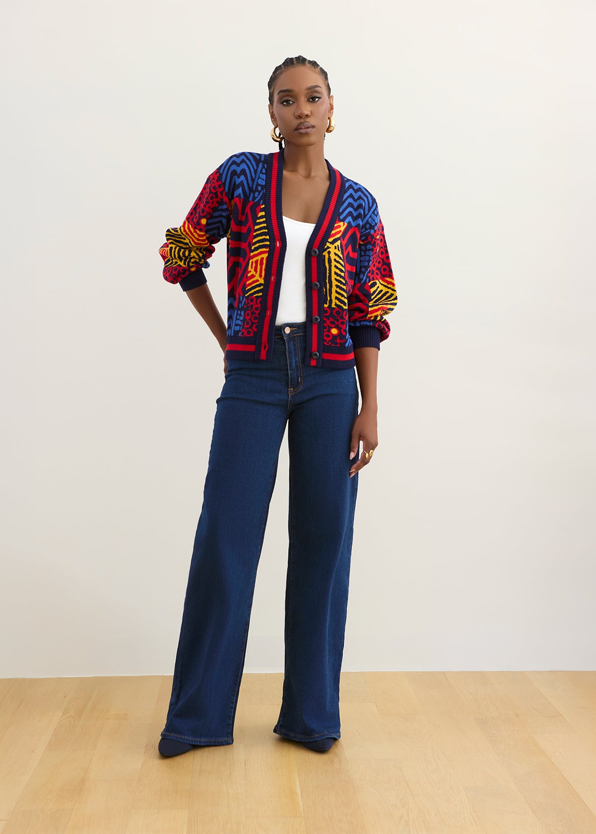 A model stands in a studio wearing a colorful patterned cardigan with blue wide-leg jeans, a white top, and gold hoop earrings against a plain off-white wall and light wood floor.