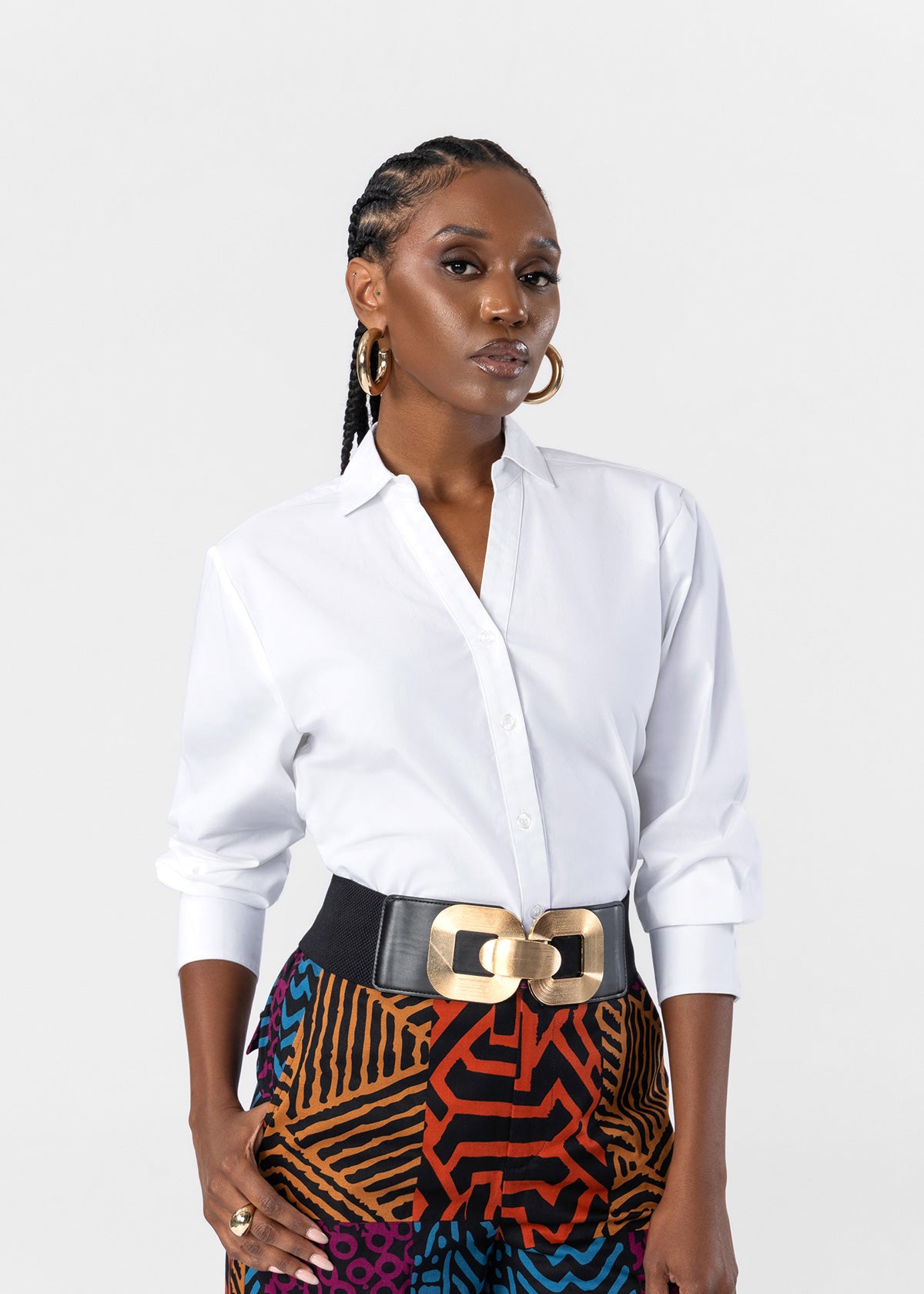 A woman modeling a white long-sleeve button-up shirt tucked into a black belt with a large gold buckle, paired with a colorful patterned skirt, against a light gray studio background.