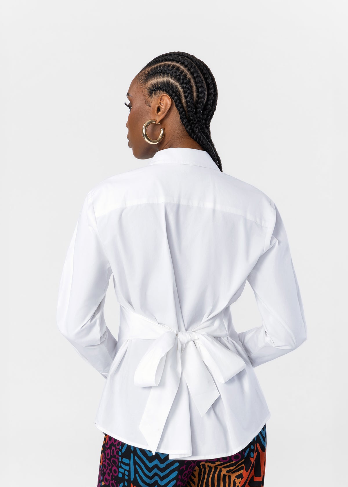 A woman shown from the back wearing a white long-sleeve collared blouse with a tied waist detail, paired with a colorful patterned skirt, against a plain light gray studio background.