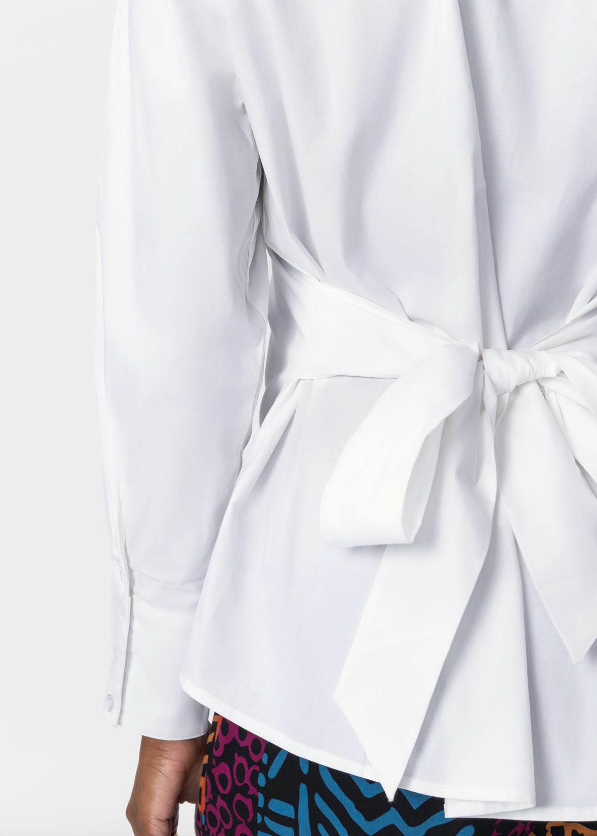 Close-up of a white long-sleeve wrap-style blouse tied at the waist, shown on a model with a colorful patterned bottom visible at the hem.