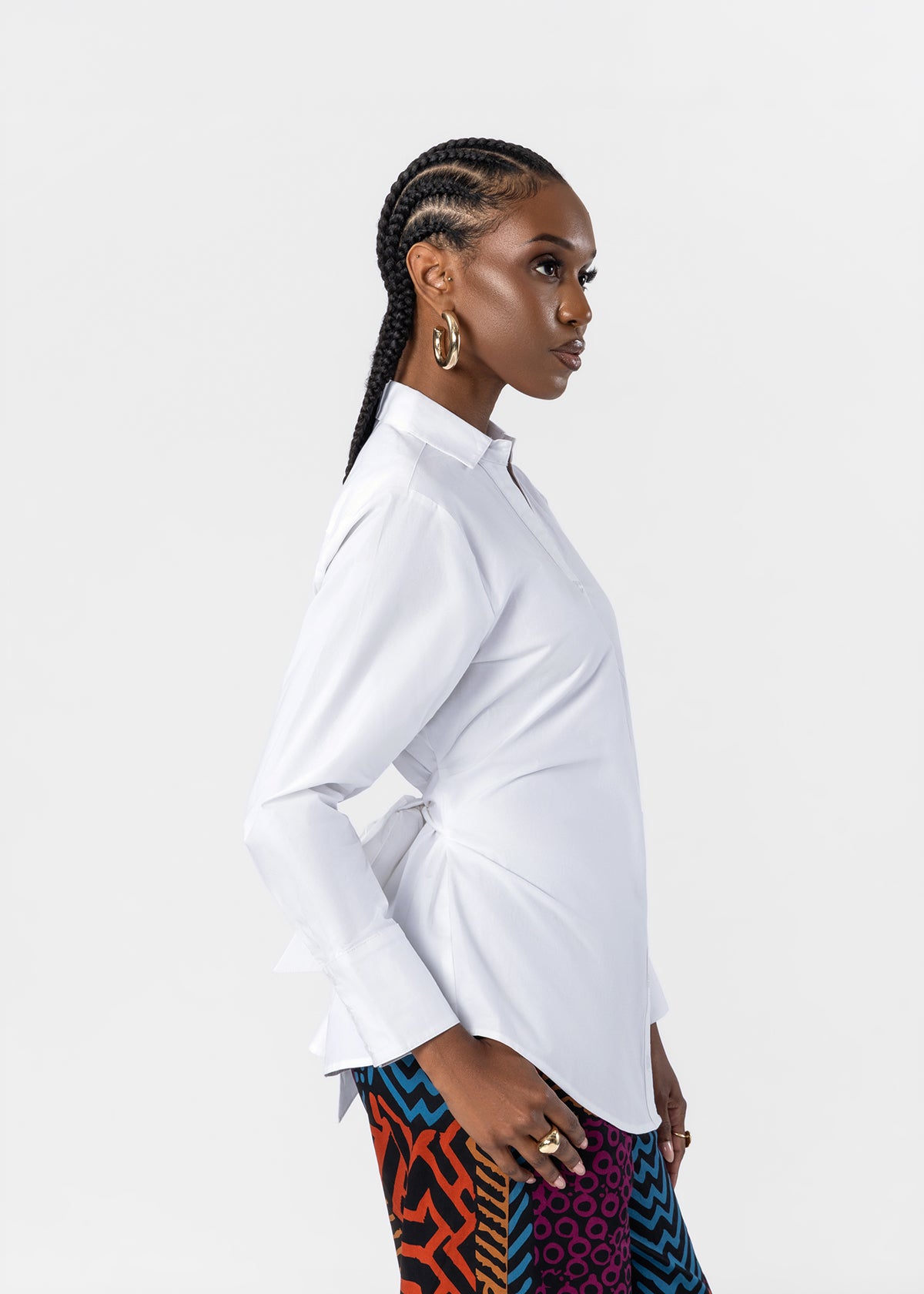 A woman in profile wearing a white long-sleeve button-up shirt and colorful patterned pants, photographed against a clean light gray studio background.