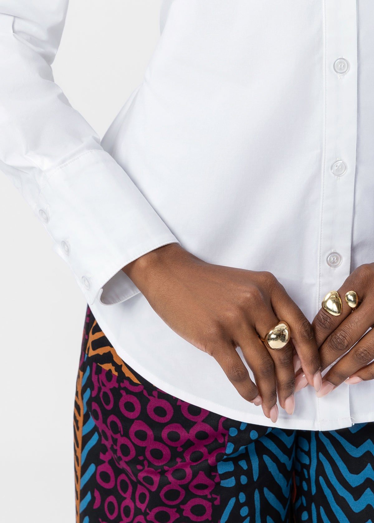 A close-up of a person wearing a white long-sleeve button-up shirt and colorful patterned pants, with gold rings on their fingers, against a light gray background.