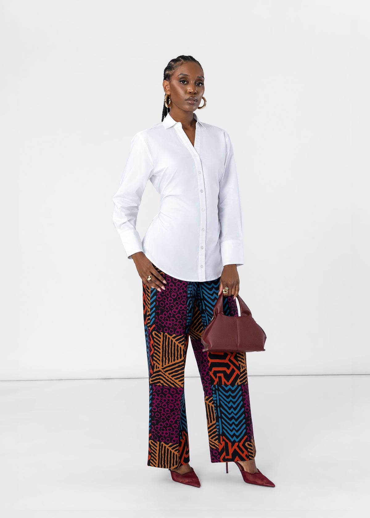 A model stands against a white studio background wearing a white button-up shirt, wide-leg pants with a vibrant purple, orange, and blue geometric pattern, and burgundy pointed-toe heels, holding a matching burgundy handbag.