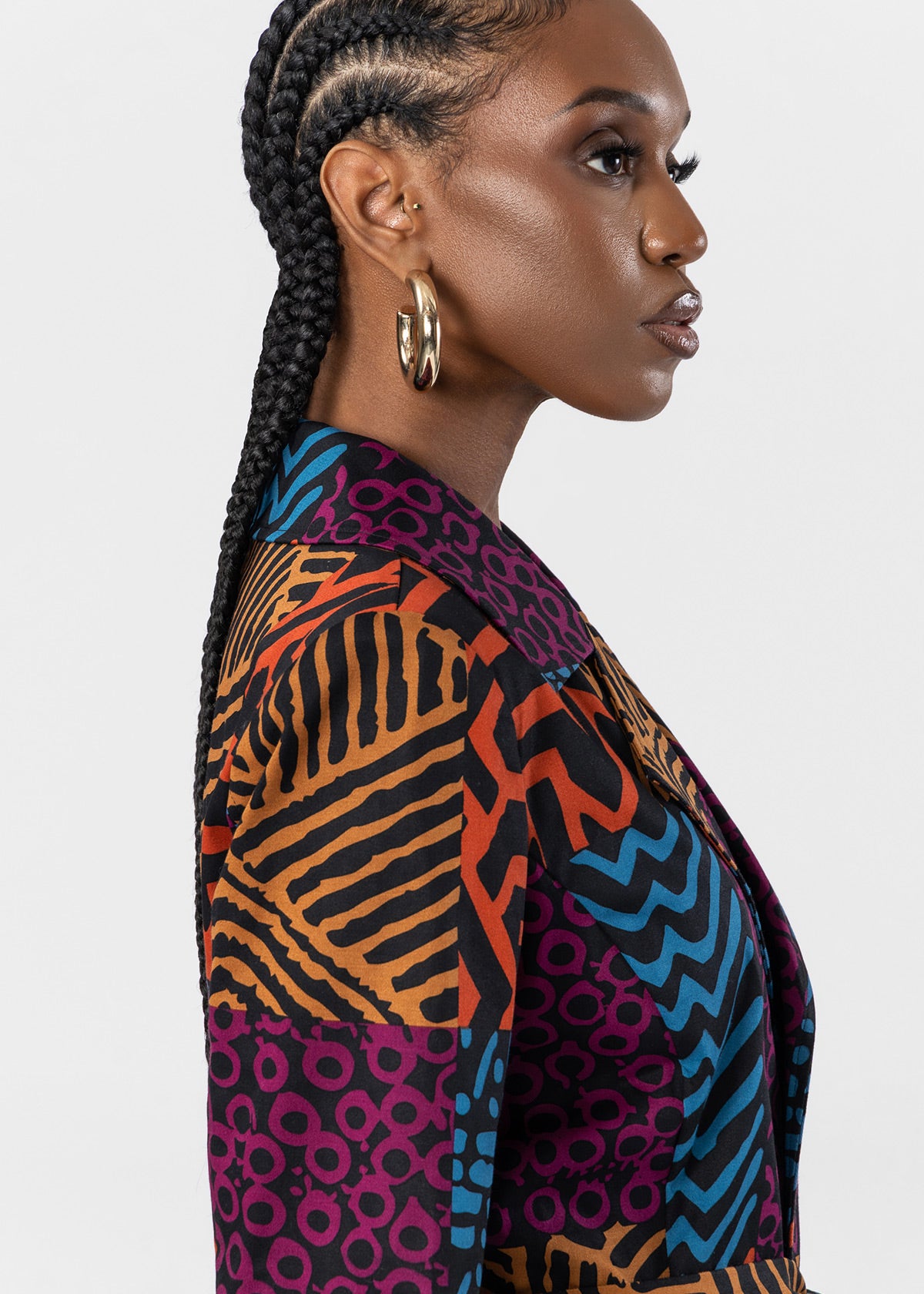 A woman in profile wearing a colorful patterned blazer with geometric and zebra-like prints in orange, blue, purple, black, and beige, paired with gold hoop earrings, against a light gray studio background.