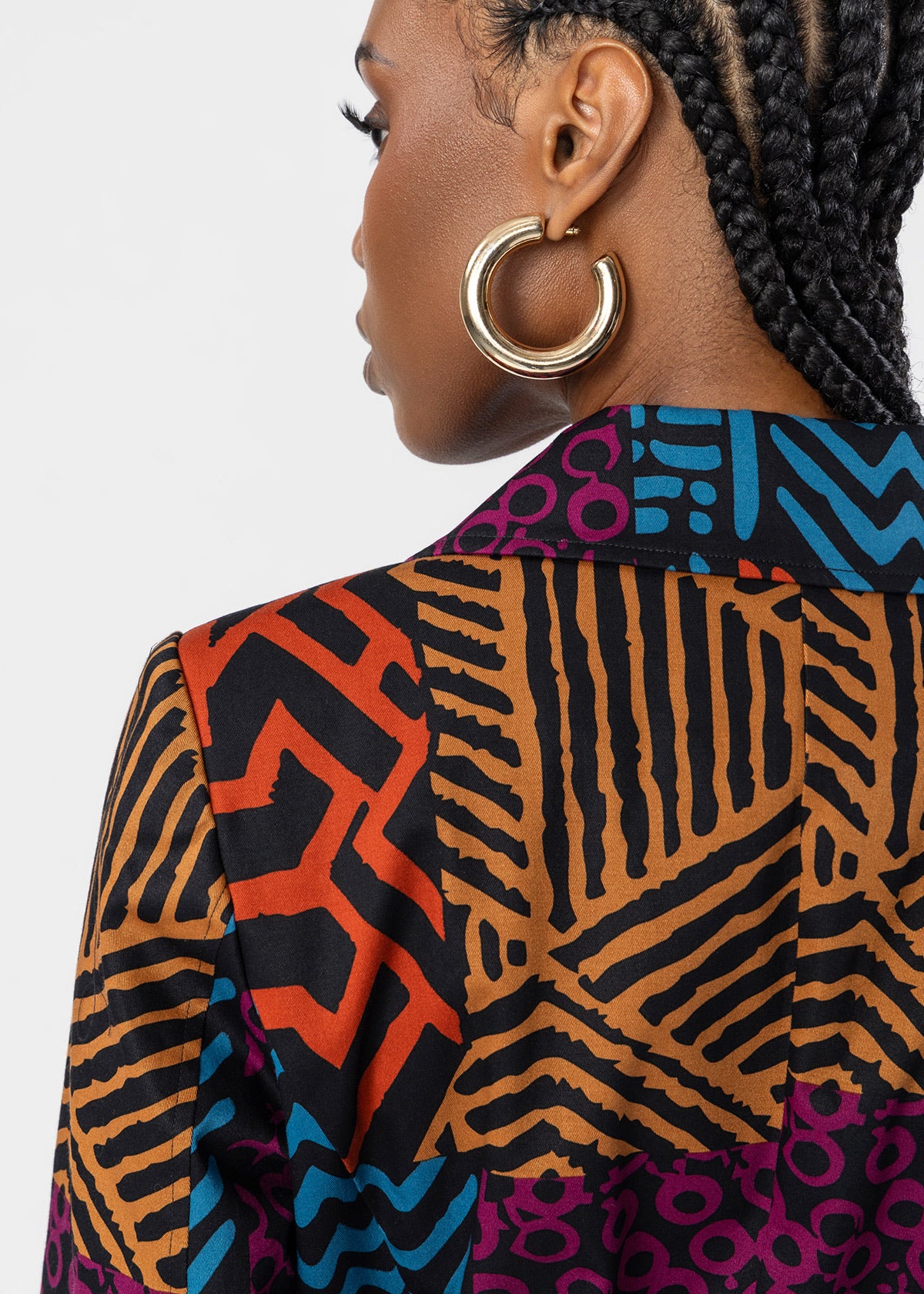Close-up side view of a person wearing a colorful patterned top with black, orange, blue, and purple geometric and zebra-like prints, paired with large gold hoop earrings, against a light gray background.