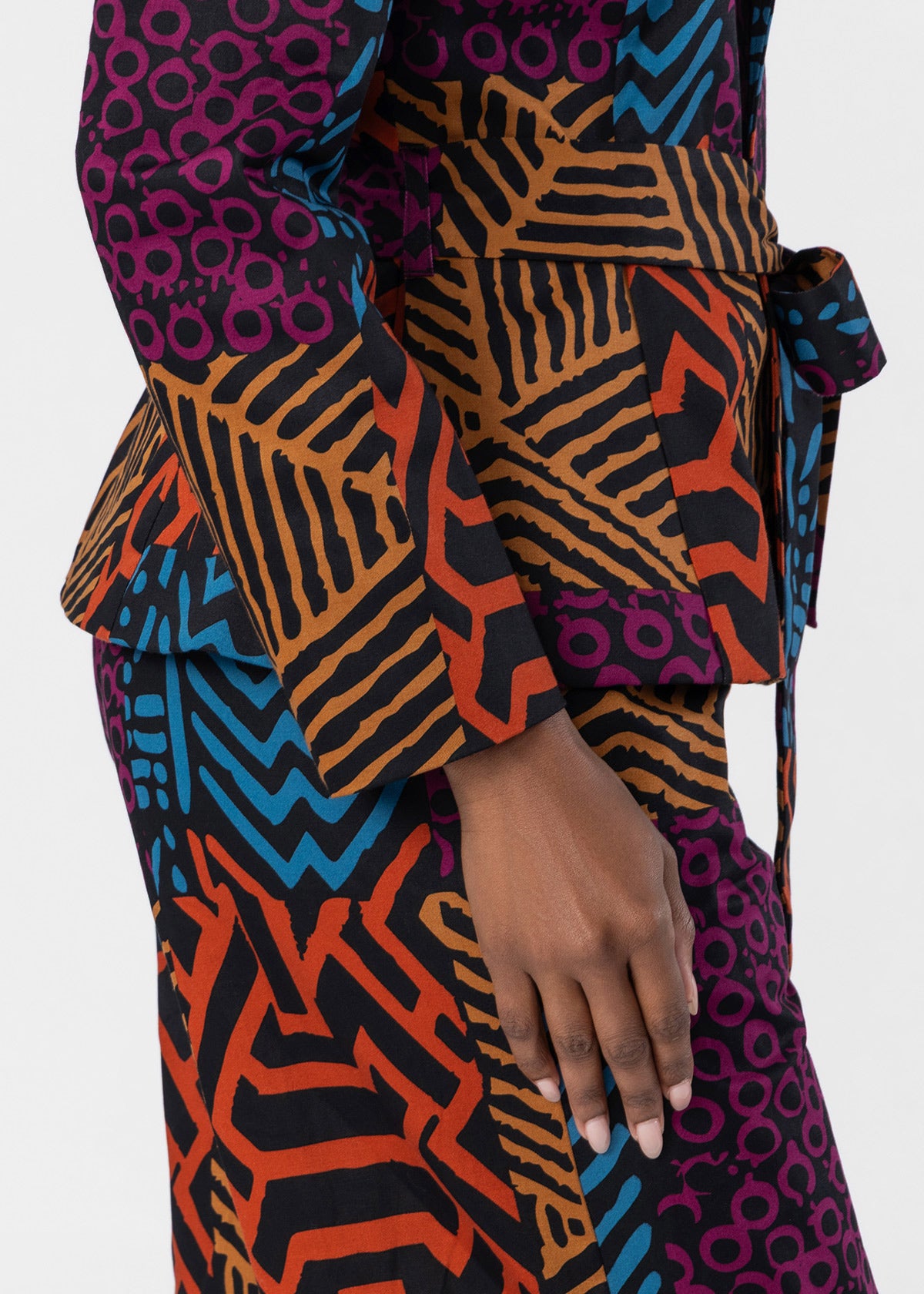 A close-up of a person wearing a matching patterned wrap-style top and skirt set. The fabric features bold geometric and abstract prints in orange, blue, magenta, black, and brown on a light gray studio background.