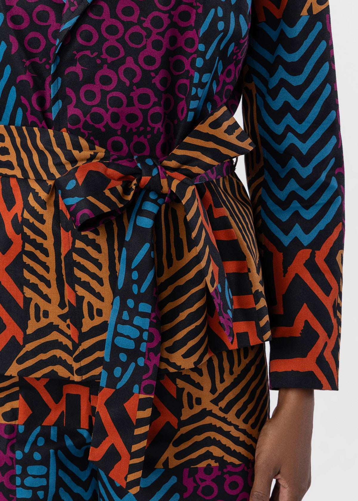 A close-up of a multicolor patterned wrap-style garment with a tie belt, featuring abstract geometric and zebra-like prints in orange, blue, black, and magenta. A hand is partially visible at the lower right.
