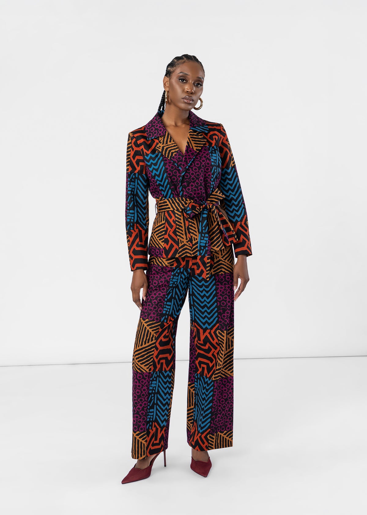 A model stands against a white studio background wearing a long-sleeve, belted jumpsuit with a bold multicolor geometric and tribal-inspired print in purple, orange, blue, and black, paired with dark red pointed-toe heels.