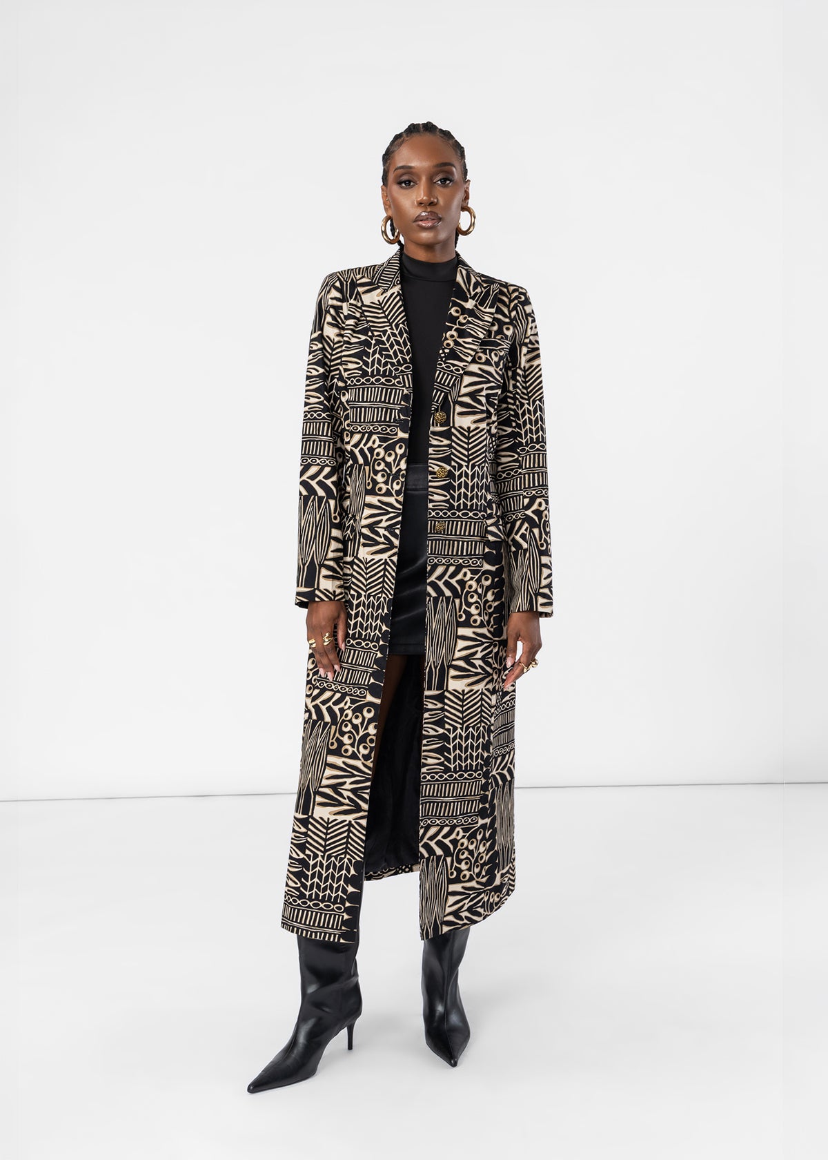 A model stands against a white studio background wearing a long black and beige geometric patterned coat over a black turtleneck and black pointed-toe heeled boots.