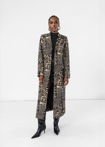 A model stands against a white studio background wearing a long black and beige geometric patterned coat over a black turtleneck and black pointed-toe heeled boots.