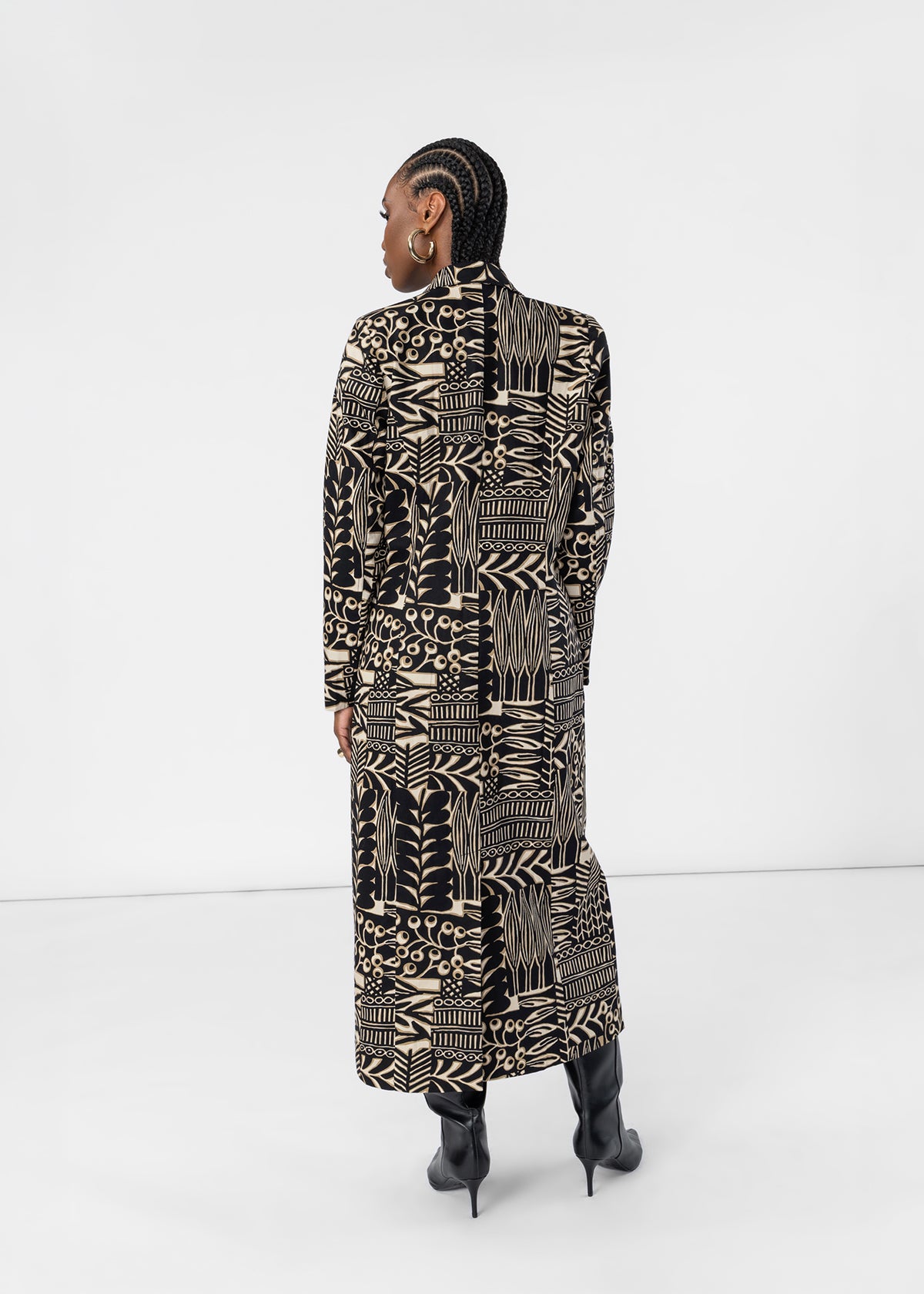 A model shown from the back wearing a long-sleeved, high-neck maxi coat with a bold black and beige geometric tribal pattern, paired with black pointed-toe stiletto boots against a white studio background.