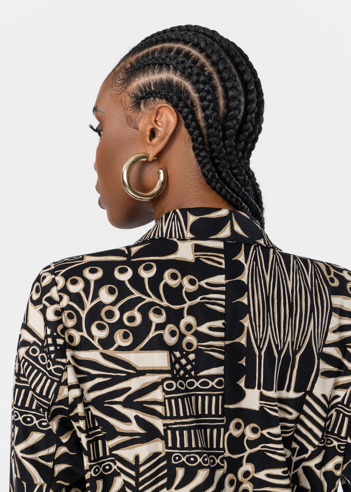 A woman shown from the back wearing a black and beige patterned shirt and large gold hoop earrings, against a light gray studio background.