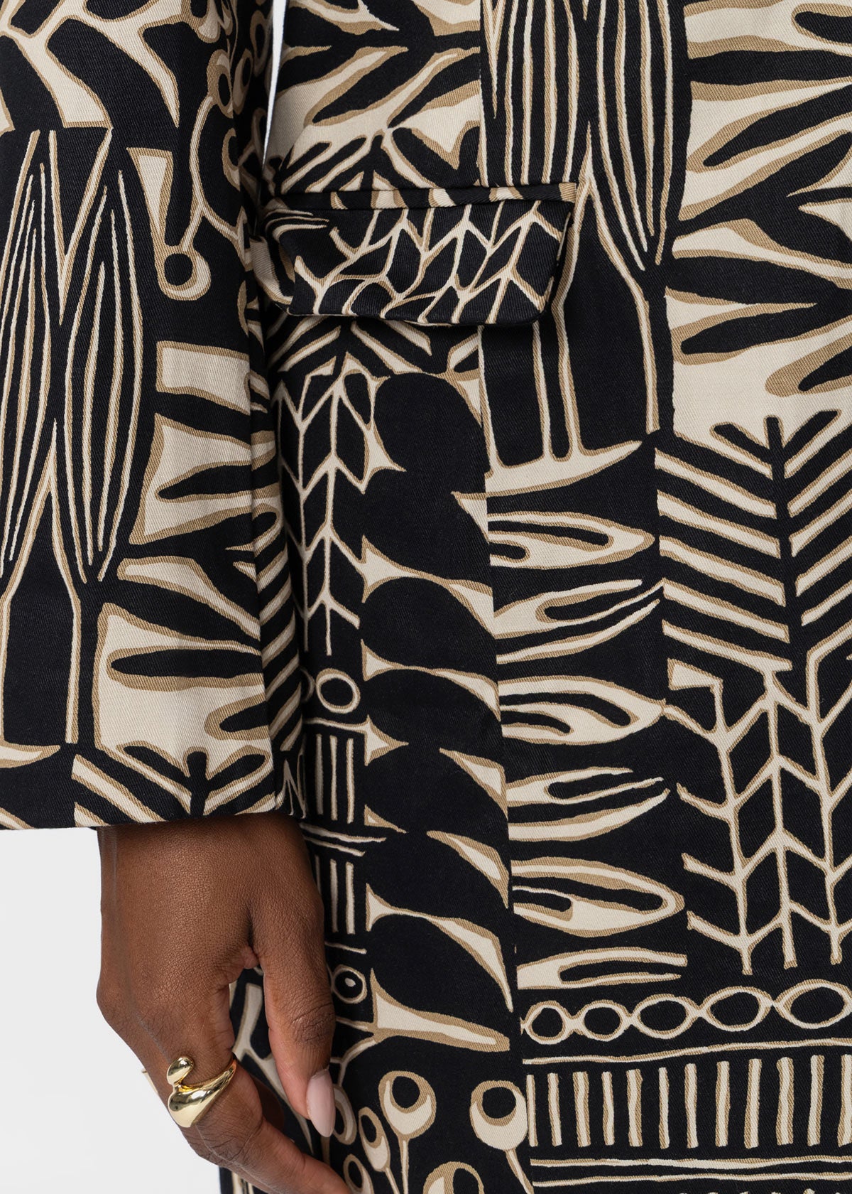 Close-up of a person wearing a belted wrap coat or jacket with a bold black and beige botanical block-print pattern. A hand wearing a gold ring is visible at the bottom left.